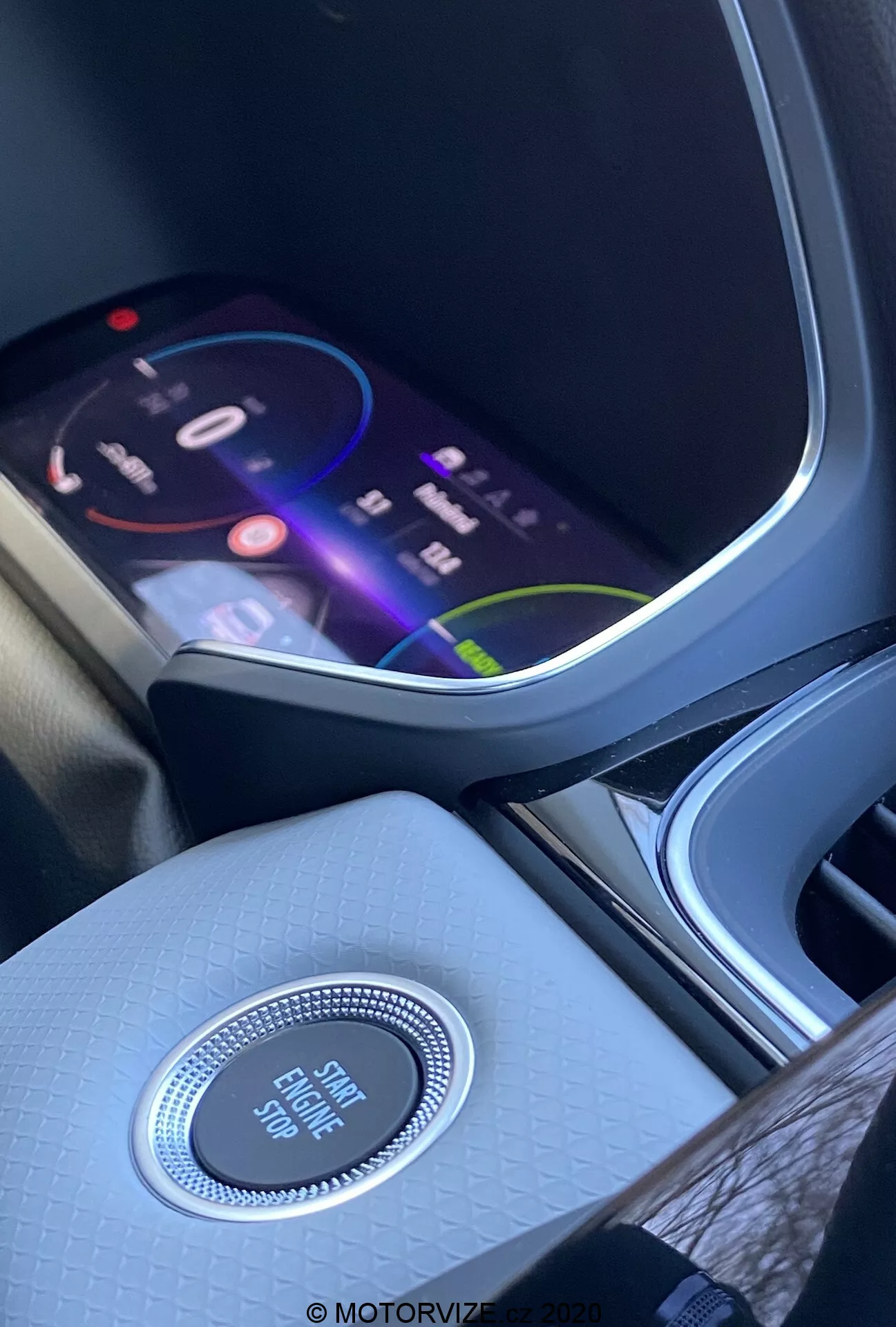 A Renault Captur 2020 belső tere, ahol a középkonzolra és a műszerfal részére helyezik a hangsúlyt az első ülés perspektívájából. Az élénk infotainment képernyő színes felületet mutat, ami fejlett multimédiás funkciókat sugall, beleértve a navigációt, médiatartalmak lejátszását és csatlakozási lehetőségeket is. A képernyő alatt található egy kör alakú, krómozott vezérlőgomb, amely 'Engine Start Stop' felirattal van ellátva, jelezve a kulcs nélküli indítási rendszer jelenlétét. A középkonzol textúrázott, kiváló minőségű anyagból készült, amely fokozza a belső hangulatot. Balra részben látható egy pohártartó vagy tárolórekesz.