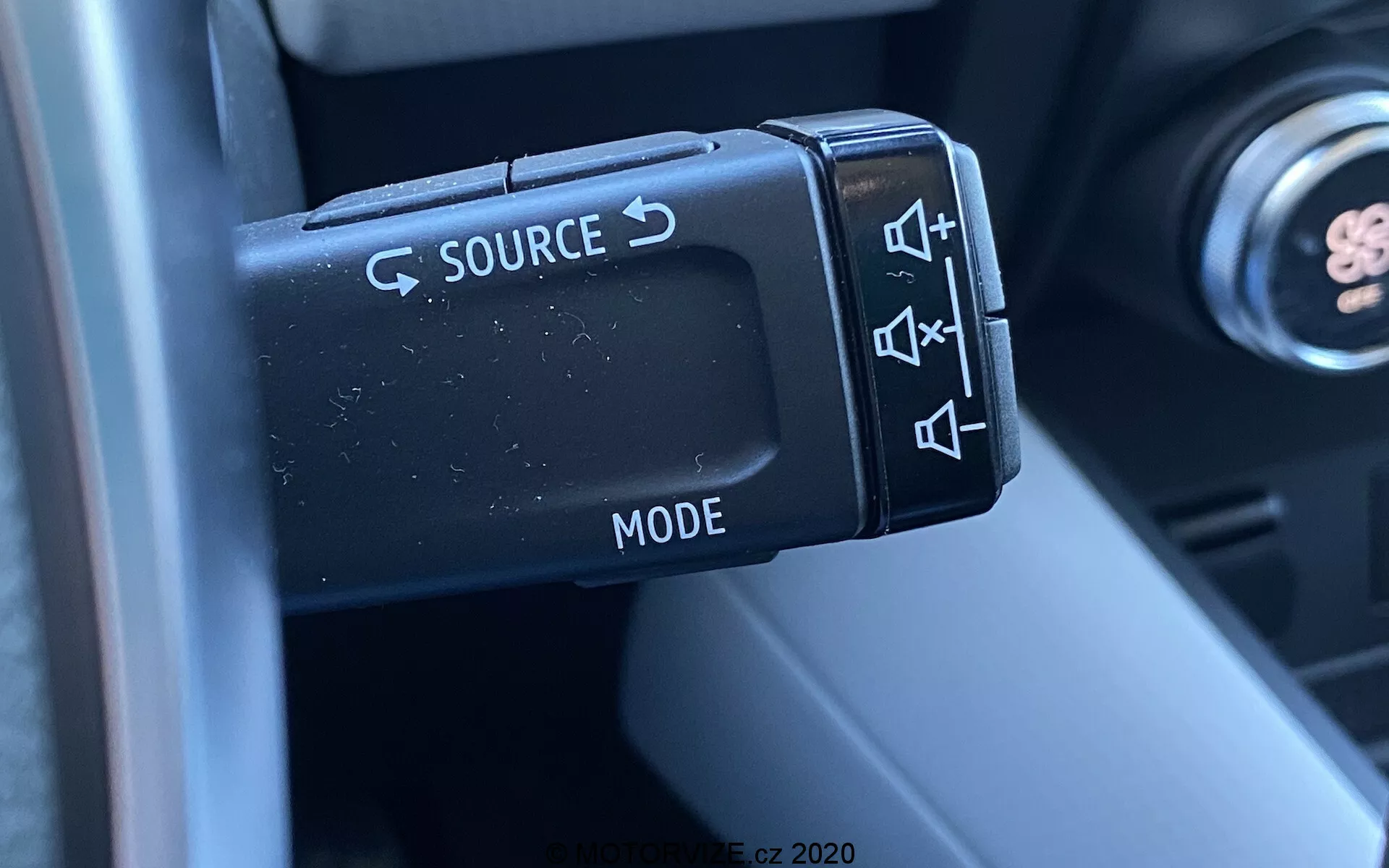 Közelkép a 2020-as Renault Captur kormánykeréken elhelyezett audio vezérlőgombjairól, a bal oldali vezérlőkarra összpontosítva, amely SOURCE cseréhez, hangerőszabályzáshoz, zeneszám ugráshoz és MODE kiválasztáshoz szolgáló gombokat tartalmaz. A fotó a matt felületű műanyag gombok és azok ergonómiai kialakításának részleteit ábrázolja, miközben a kormányoszlop és az autó belső részét elmosódott háttérként mutatja be, kiemelve a vezető számára nyújtott funkcionalitást és kényelmet.