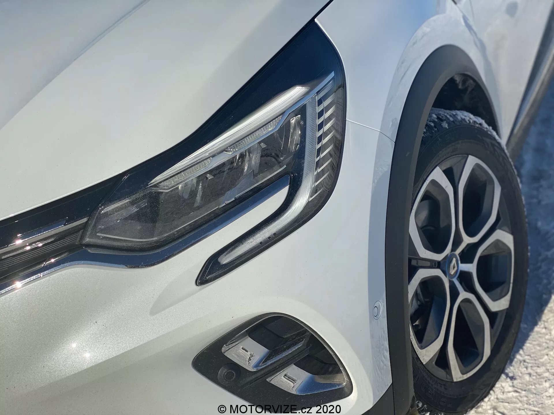 Közelkép a 2020-as Renault Captur első bal negyedéről, elegáns LED-es fényszórókkal, stílusos többrészes alumínium kerékkel, dinamikus első lökhárítóval légbeömlő nyílásokkal és fényes króm díszítésekkel a fényes fehér karosszérián.