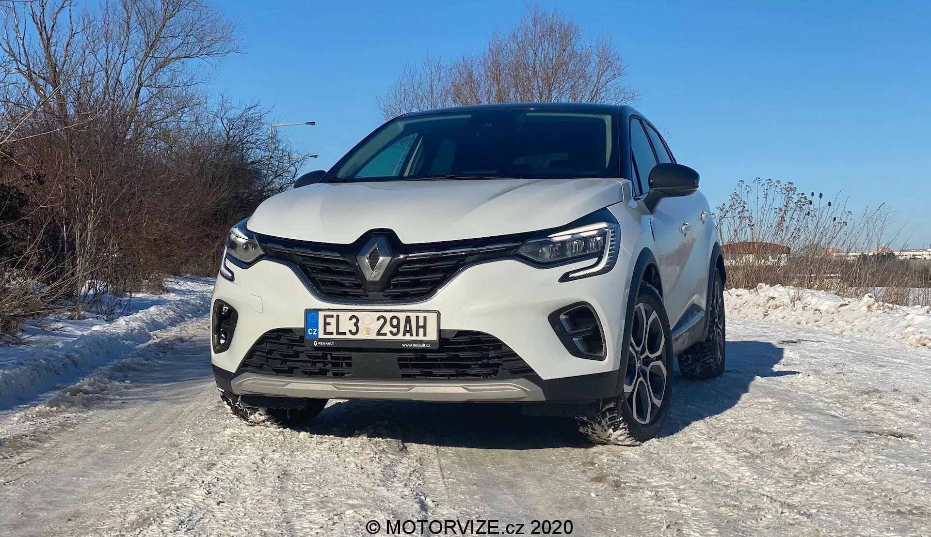 Egy fehér 2020-as Renault Captur elülső nézete, enyhe bal oldali szögből fényképezve. A modern jármű különleges Renault hűtőrács dizájnt mutat nagy központi logóval, éles szögletes vonalakkal és formás kontúrokkal. Jól látható az egész hűtőrács, a LED fényszórók és az éles nappali menetfények. A sportos lökhárító feltűnő oldalsó légbeömlőkkel és valószínűleg hűtésre szolgáló középső résszel rendelkezik. A motorháztetőn domború vonalak találhatók, amelyek izmos megjelenést kölcsönöznek a járműnek. A havas felületen látható az első kerekek és a sokküllős könnyűfém felnik egy része, ami téli körülményekre utal. Az európai rendszámtábla és a fényes festék, amely visszatükrözi a napfényt, egy tiszta, napsütéses napot sugall. A 'E' rendszámtáblából kiindulva feltételezhetően haladó vezetőtámogató rendszerek, LED világítás és hibrid/elektromos hajtáslánc lehetnek.