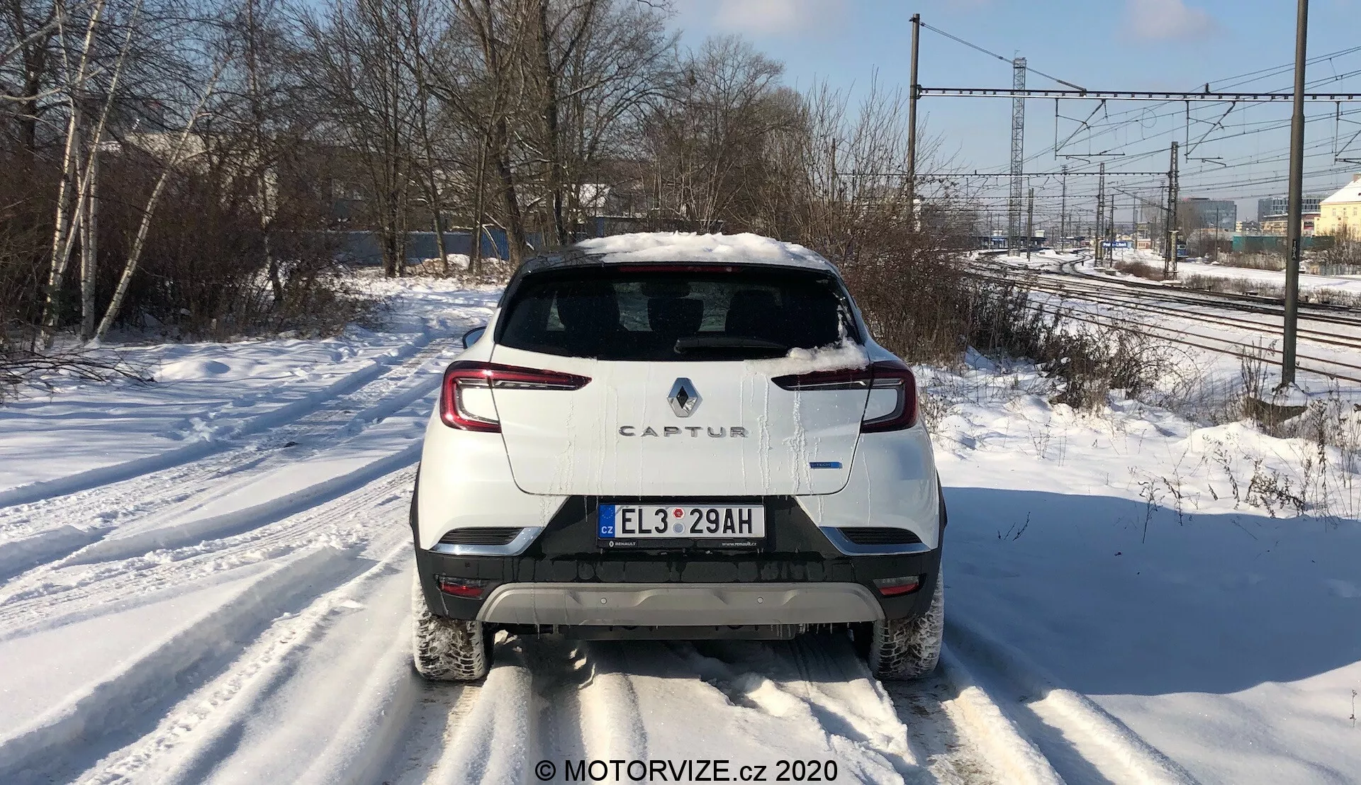 Középső hátsó nézet egy 2020-as Renault Captur-ről egy havas úton. A kép kiemeli a jellegzetes hátsó kialakítást, beleértve a kéttónusú színsémát fehér testtel és kontrasztos tetővel, modern hátsó LED világítást, európai stílusú rendszámtáblát, hátsó ablaktörlőt és egyetlen kipufogócsövet a jobb oldalon. A fotón áramvezetékek és fasorok láthatók a háttérben, ami elővárosi vagy félvidéki környezetre utal.