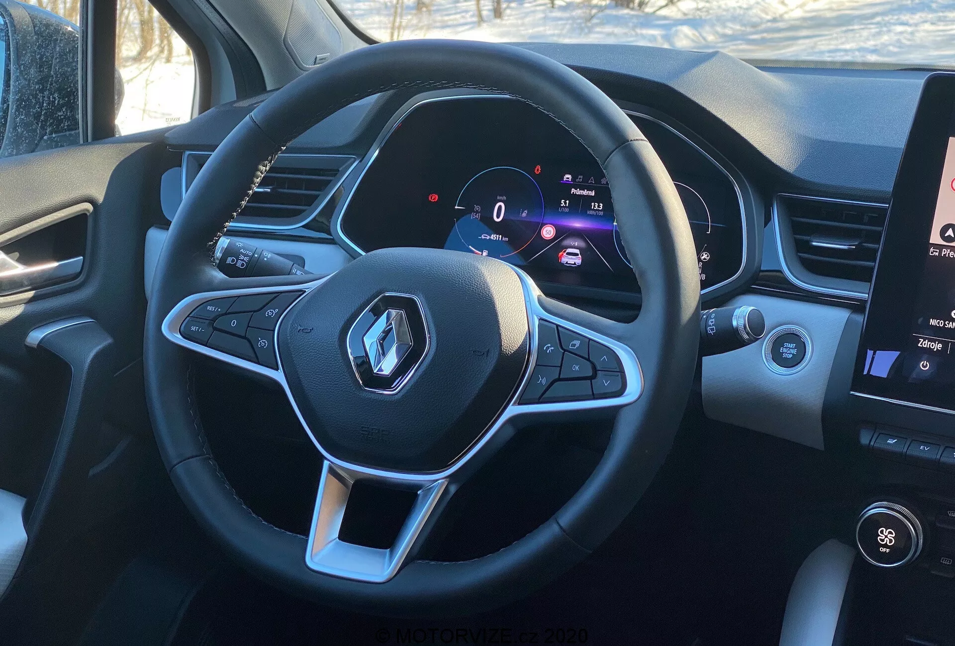 A 2020-as Renault Captur belső nézete a vezető szemszögéből, hangsúlyozva a sportos lapos aljú multifunkcionális kormánykereket Renault logóval és az audió és tempomat funkciók vezérlő gombjait. A digitális műszerfal 0 km/h-os álló sebességet és a fordulatszámmérőt mutatja, jelezve a modern technológiát és a testreszabható vezetői információkat. A jobb oldalon látható az infotainment rendszer függőleges érintőképernyőjének egy része, amely a hőmérséklet-szabályozást és a média információkat jeleníti meg, nappali fényviszonyok között, valósághű vezetői üléspozíciót biztosítva.