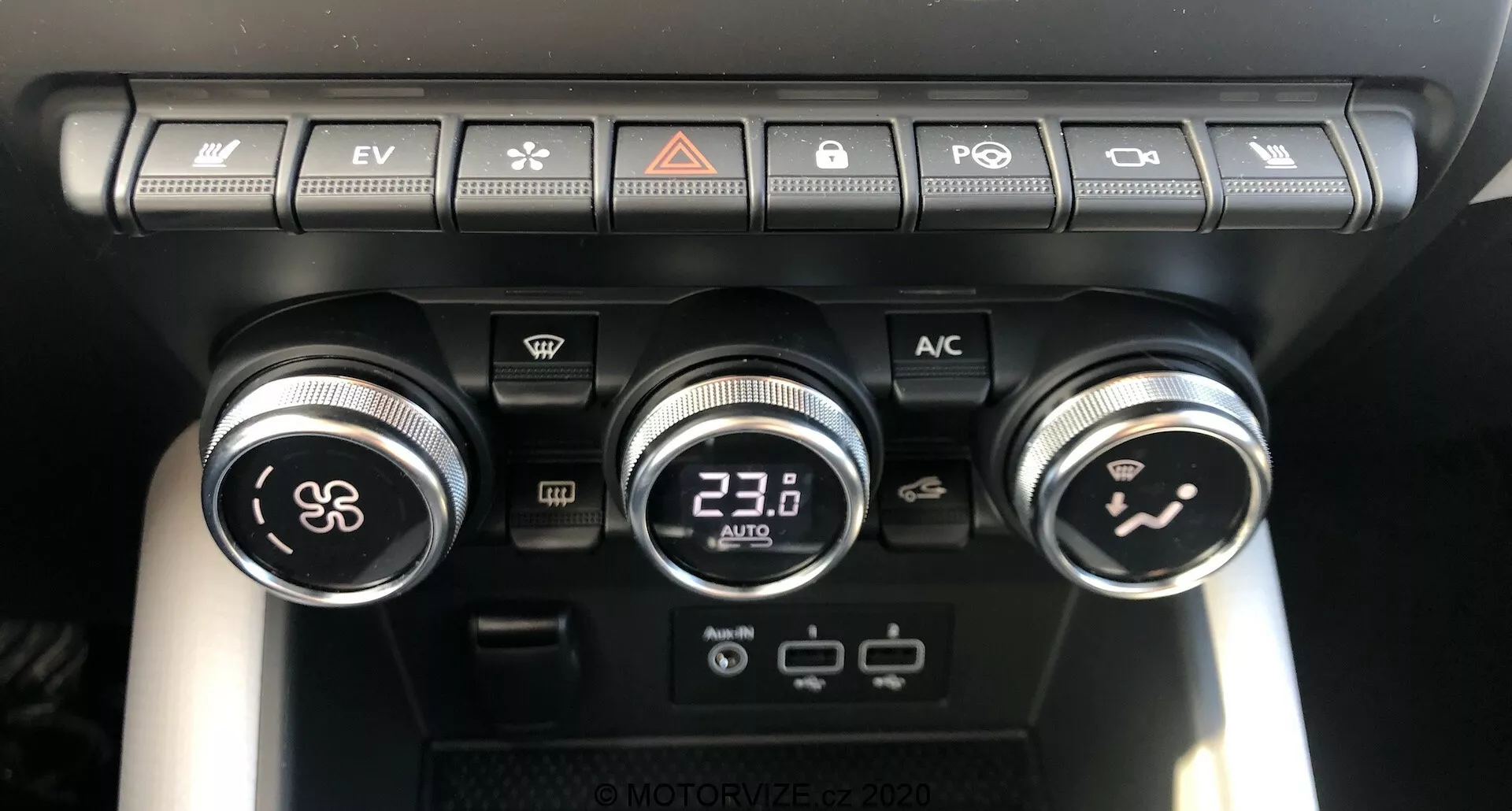 Közelkép a Renault Captur 2020 klímavezérlő paneljéről a jármű belsejében, amely három tárcsát és több gombot mutat. A bal oldali tárcsa szabályozza a ventilátor sebességét, a középső tárcsa digitális kijelzővel 23,0°C-ra van állítva, a jobb oldali tárcsa pedig az utastér légáramlását irányítja. A tárcsák felett egy sor gomb található, amelyek között van a levegő újrakeringetése, a szélvédő párátlanítása, a hátsó ablak párátlanítása, a hőmérsékletszinkronizáló, az automatikus üzemmód kapcsoló és az 