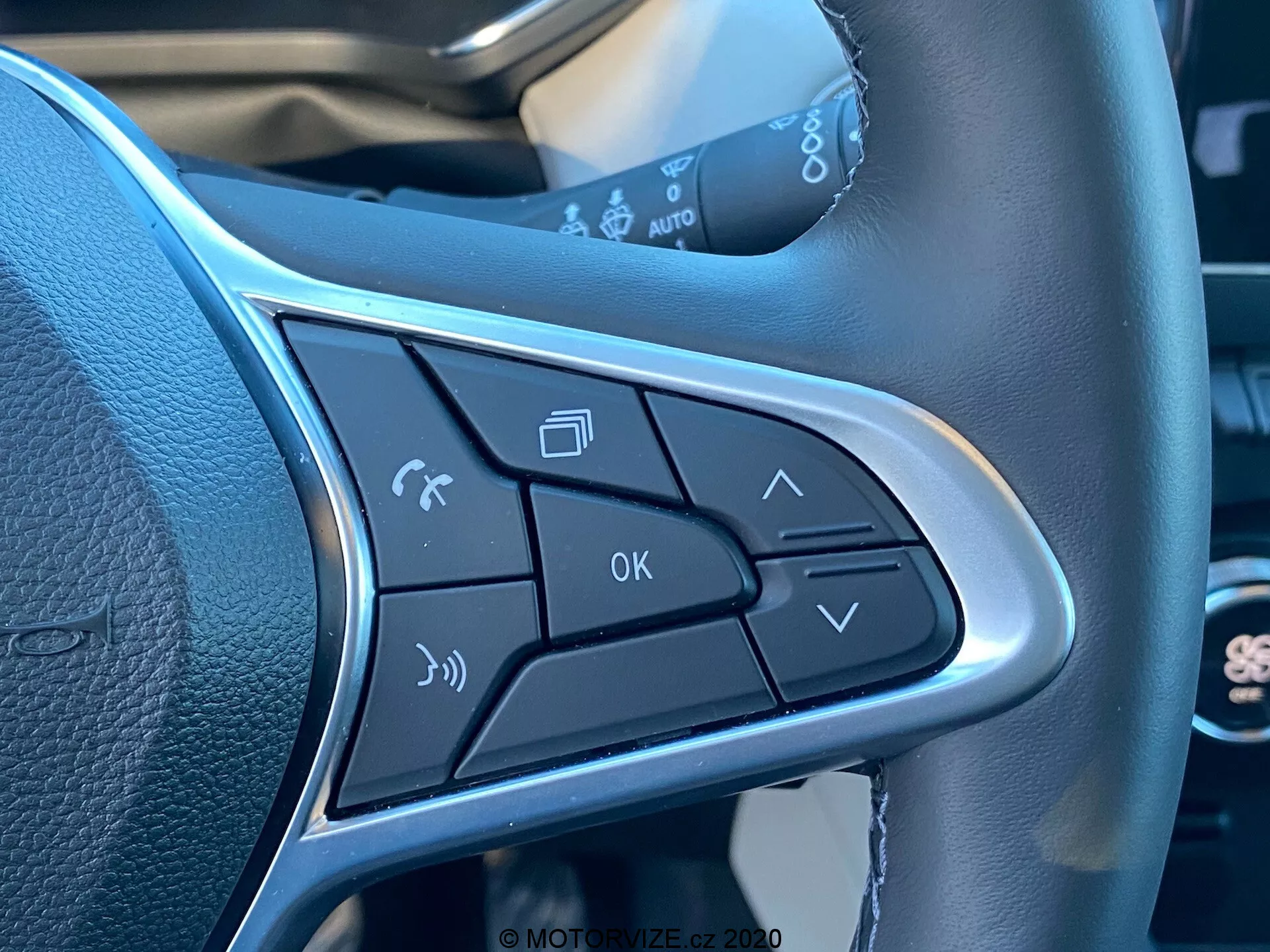 Közelkép a Renault Captur 2020 kormánykerék küllőjén található vezérlőgombokról. Funkciók közé tartozik a hangparancs aktiválása, telefonkezelés, hangerőszabályzók, OK gomb és menünavigációs nyilak. Az elegáns kialakítás része az ezüst szegély, amely kiemeli a gombokat, hangsúlyozva a kéz nélküli működést és a kényelmet.
