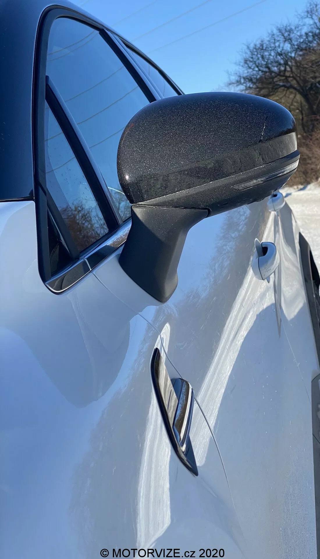 Részletes közelkép egy fehér 2020-as Renault Captur oldalsó tükréről, ajtajáról és első negyedpaneljéről. Az oldalról közvetlenül az oldalsó tükör előtt felfelé irányított szögből készült fotó kiemeli az oldalsó tükör modern, aerodinamikailag hatékony kialakítását fekete texturált alsó résszel a tartósság érdekében. Az áramvonalas tartó simán illeszkedik az ajtókeretbe. Az ajtó süllyesztett kilincse és az azon végigfutó jellegzetes vonal karcsú, sportos dizájnt sugall. A fényszóró egy része és az ablakban visszatükröződő tiszta ég erősíti ezt a dinamikus megjelenítést.