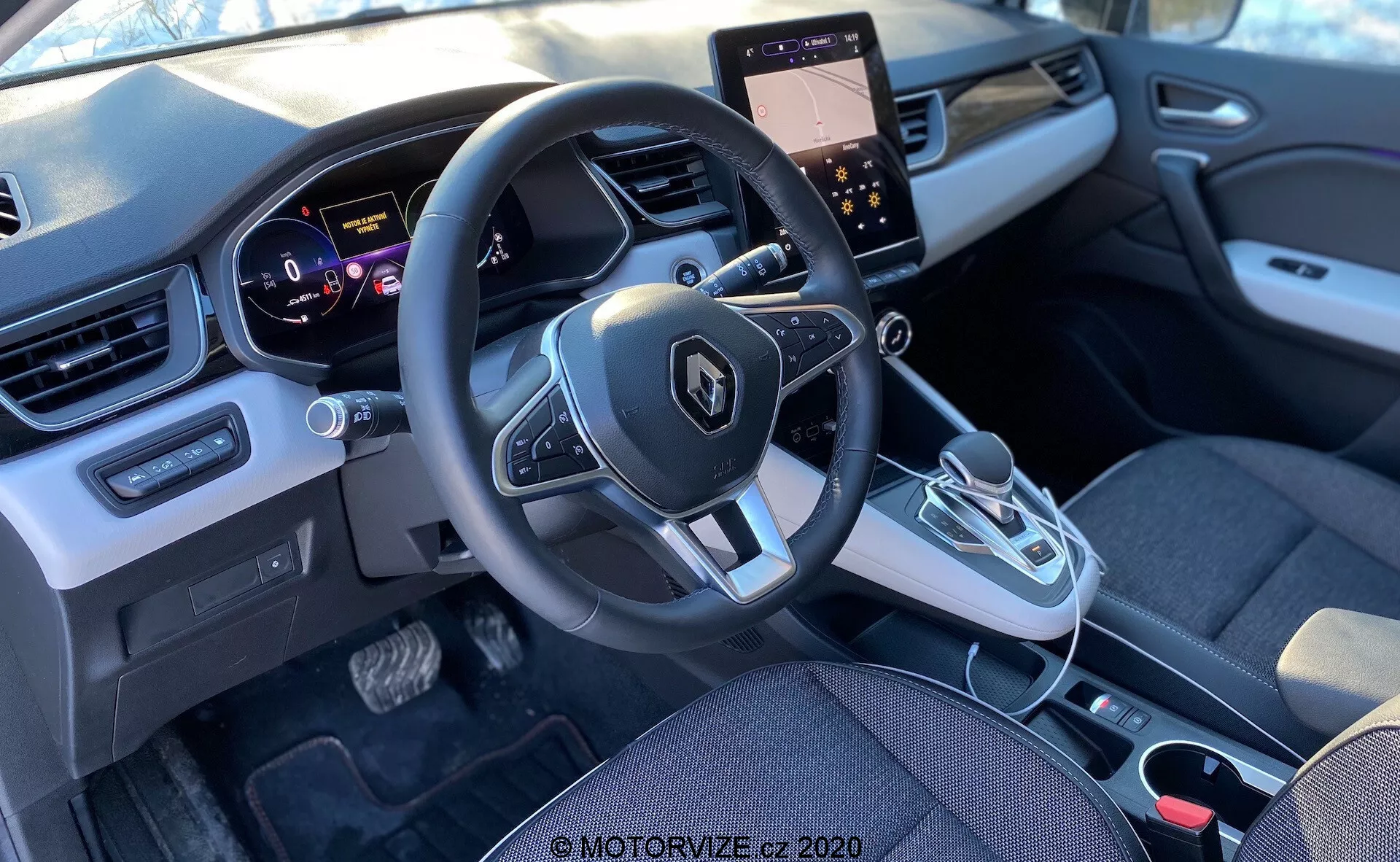 2020-as Renault Captur belső nézete a hátsó ülésből, az első kabinra összpontosítva, beleértve a műszerfalat, a kormánykereket beépített vezérlőkkel, a műszercsoportot, a középkonzolt érintőképernyős kijelzővel és klímavezérléssel, az automatikus sebességváltó kart, valamint a szövet- és műanyag elemek modern keverékét.