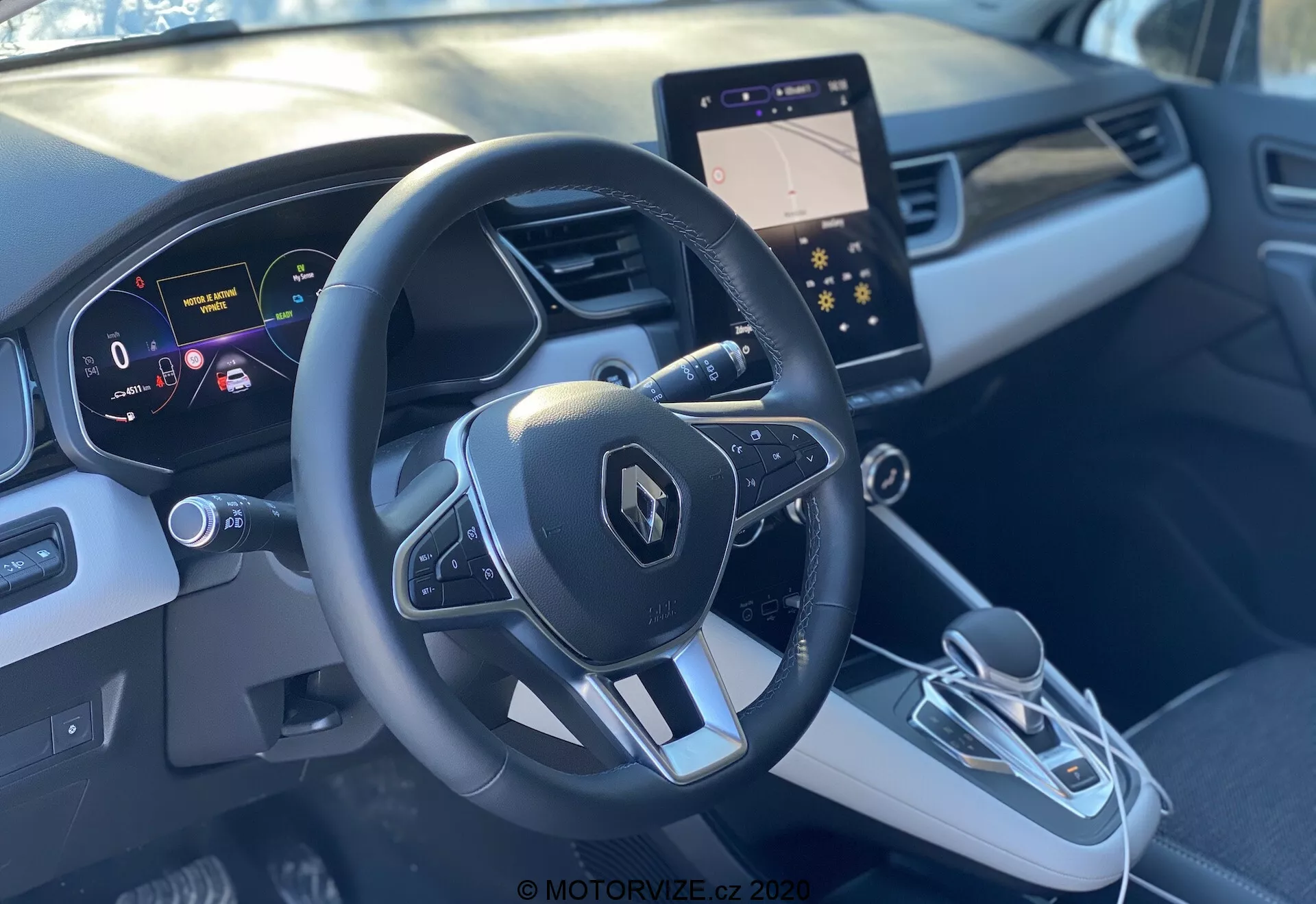 A Renault Captur 2020 belső nézete az első utasülésből, bemutatva a sportos, háromküllős kormánykereket Renault emblémával és multifunkciós gombokkal, a digitális műszercsoportot tiszta, nagy felbontású kijelzővel, amely a sebességet és a navigációs segédeszközöket mutatja, valamint az elegáns középkonzolt. A középkonzol modern infotainment képernyővel, elektronikus váltóval és elektronikus rögzítőfék gombbal rendelkezik. A kéttónusú szürke belső tér kontúros kialakítással és díszítő elemekkel prémium érzést biztosít.