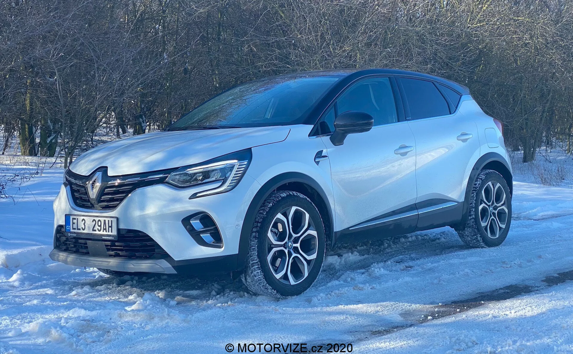 Alacsony szögből készült kép a 2020-as Renault Captur SUV-ról havas felületen, amely az elülső negyed profilját mutatja. A ragyogó nappali fény és a tiszta égbolt kiemeli a modern dizájnelemeket, mint a prominens első hűtőrács emblémával, LED-es nappali menetfények, összetett, kétszínű könnyűfém felnik, szobrászati karosszéria kiemelkedő kerékívekkel, karosszéria színű tükrök beépített irányjelzőkkel, sötétített ablakok és süllyesztett ajtókilincsek. A fotó hangsúlyozza a jármű prémium minőségű kivitelezését és potenciális téli vezetési képességeit.