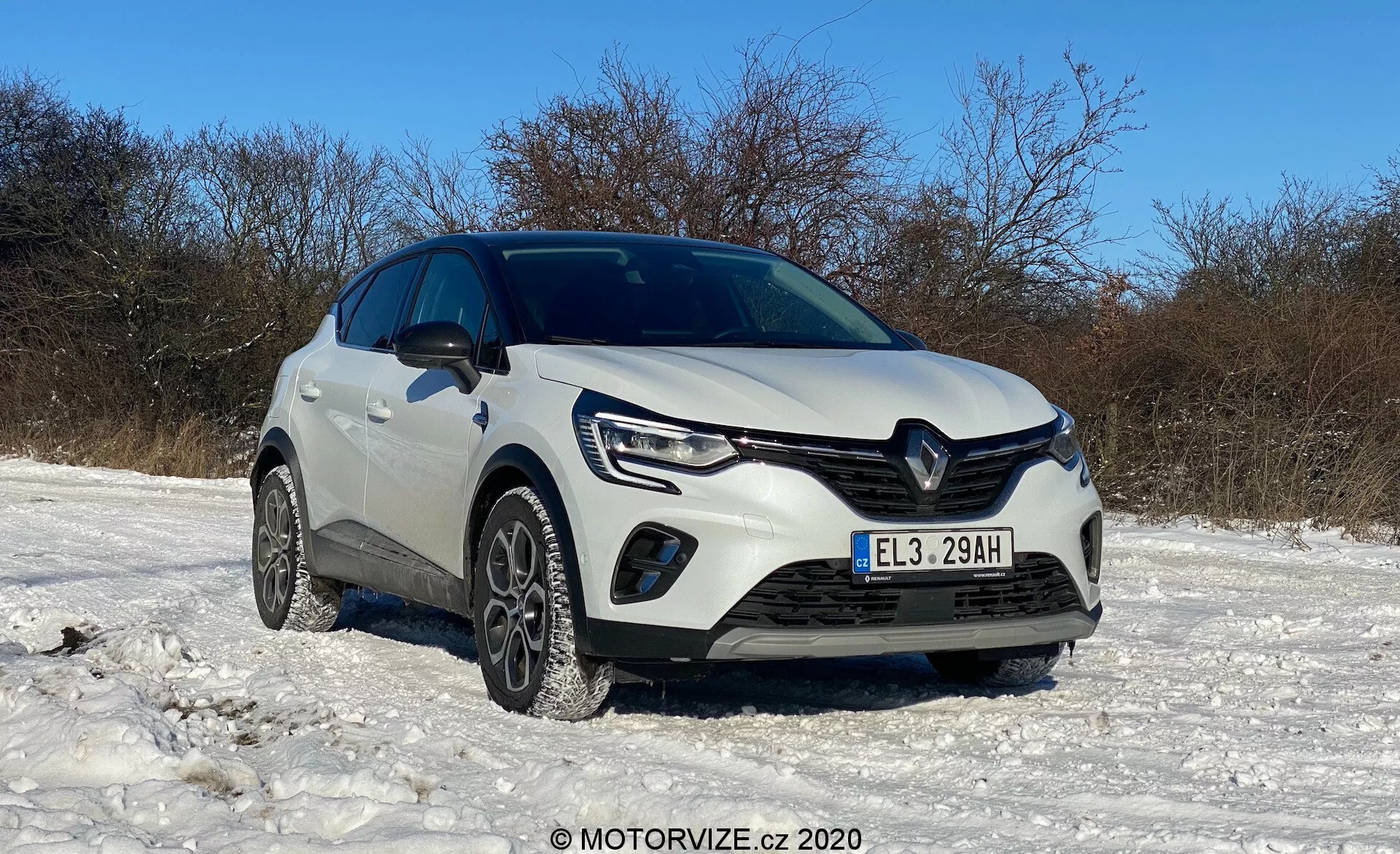 Renault Captur 2020 fehér színben, hóborította terepen parkolva, tiszta ég alatt, alacsony elülső háromnegyedes szögből bemutatva, látható első rácsozattal, LED nappali menetfényes fényszórókkal, ködfényszórókkal, első lökhárítóval, téli gumiabroncsokkal, jellegzetes Renault logóval, csiszolt motorháztető vonalakkal, megemelt hasmagassággal, esetleges parkolási asszisztens szenzorokkal, oldalsó tükrökkel és általánosan tiszta, jól karbantartott állapotban, MOTORVIZE.cz © 2020 vízjelével.