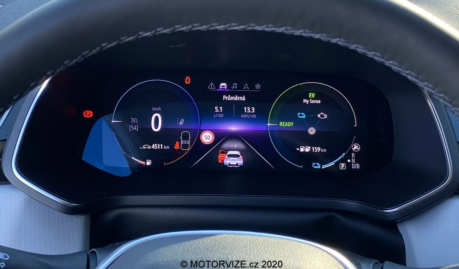 Közelkép a Renault Captur 2020 modern autó digitális műszerfal kijelzőjéről a vezető szemszögéből. A teljesen digitális, nagy felbontású, színes kijelző, amely egy texturált műszerfalba van ágyazva, látható varrással, 0 km/h sebességmérőt mutat 50 km/h sebességkorláttal, középen egy animált autó grafikát biztonsági figyelmeztetésekkel és akkumulátorjelzőkkel, egy fordulatszámmérőt az öko-barát funkciót jelképező 'My Sense' ikonnal, megtett kilométer-számlálóval, külső hőmérséklet kijelzővel, 'Park' fokozatban lévő sebesség jelzéssel, sávban tartó asszisztens ikonjaival, automatikus távolsági fényszórókkal és adaptív sebességtartó automatikával, mindezt jól szervezett elrendezésben, amely kiemeli az autóipari technológia és belső dizájn fejlettségét.