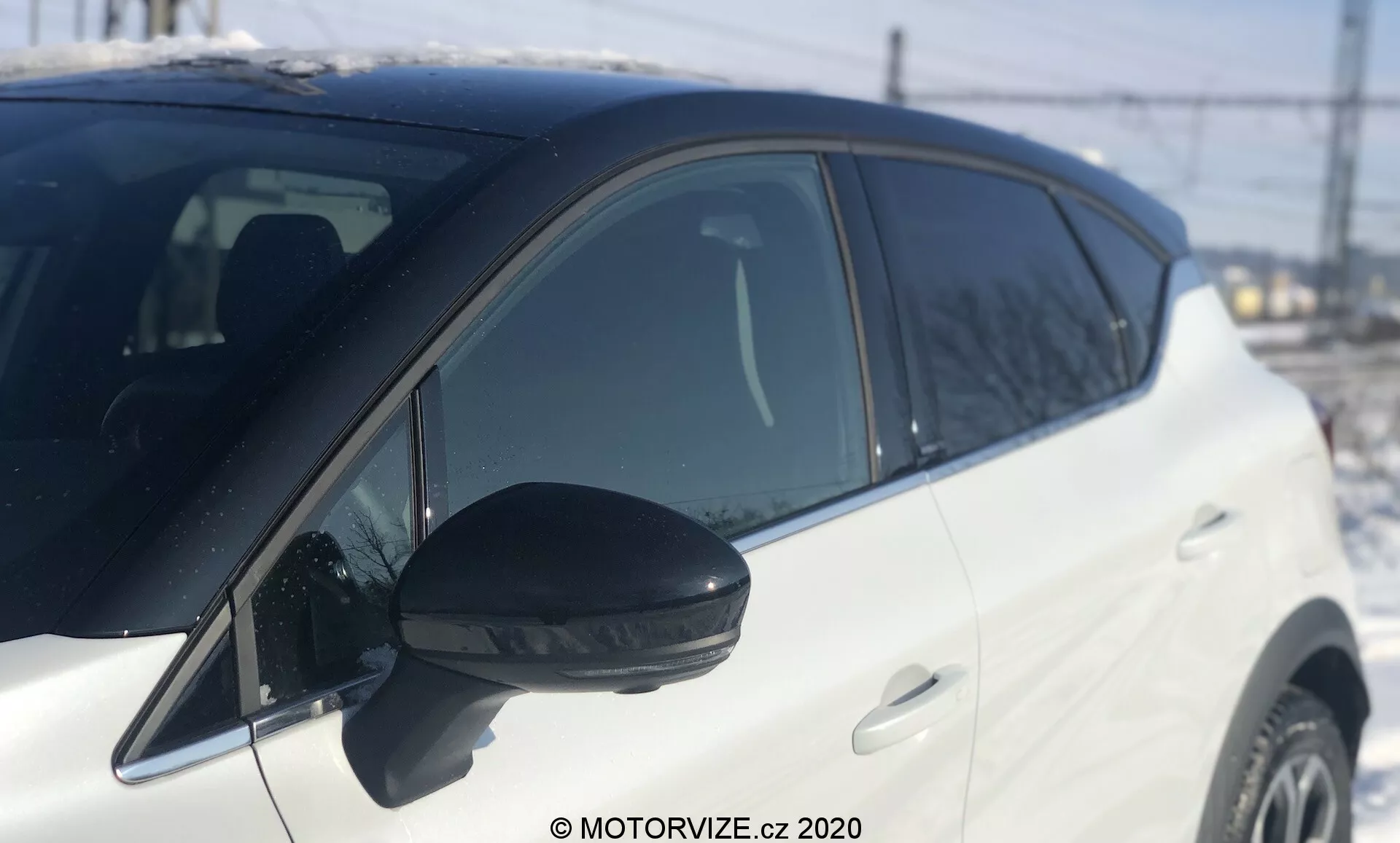 Renault Captur 2020 fehér autó közeli oldalnézet, a visszapillantó tükörtől az első kerék előtti részig. Jellegzetes fekete oldalsó tükör beépített irányjelzővel, nappali fényben, természetes napfénynél készült felvétel. Enyhén alacsony szög, amely a jármű középrészére összpontosít, sekély mélységélességgel. Elmosódott háttér, felismerhető elemekkel, mint kerítés, elektromos vezetékek és épület, amelyek kültéri, esetleg ipari vagy félvárosi környezetet sugallnak. A jármű fényes fehér festése jól visszaveri a fényt, tükrözve a jól karbantartott, modern járművek felületét. Színben egyező ajtókilincsek és emelkedő ablakvonal, amelyek dinamikus stílust idéznek.