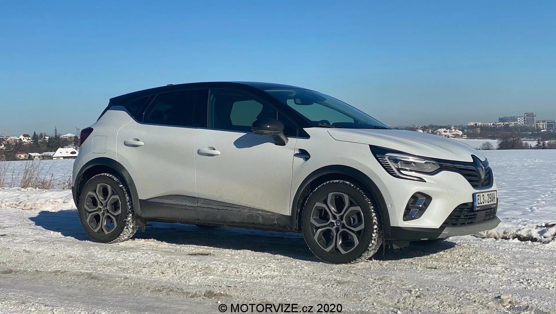 Renault Captur 2020 modern crossover SUV jéghideg, havas terepen, tiszta égbolt háttérrel. Az utasoldal néz enyhe szögben a kamerába, mutatva az autó elejét, oldalát és részben a tetejét. Jellemzők: LED nappali menetfények, gyártói emblémával integrált első hűtőrács, két tónusú világos és fekete fényezés, öt küllős könnyűfém felnik, műanyag védőburkolatok, elegáns, süllyesztett ajtókilincsek és egy hátsó spoilerrel záródó ablakvonal. Fejlett technológiát sugall, mint a LED világítás és a kulcsnélküli bejutás, hideg időjárási környezetben, közel városi területhez.