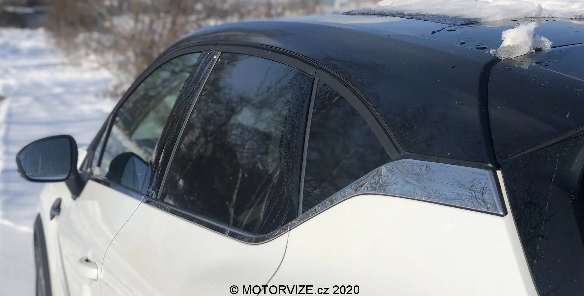 A Renault Captur 2020 oldalnézete egy téli környezetben, kiemelve a fekete-fehér, két tónusú festési sémát. A kép a B-oszlopra, a hátsó ajtó egy részére és a hátsó negyediszterületre koncentrál, kissé hátrafelé tekintve. A tetőn lévő hórétegek és az ablakon olvadó jég egy fényes napot tükröznek.