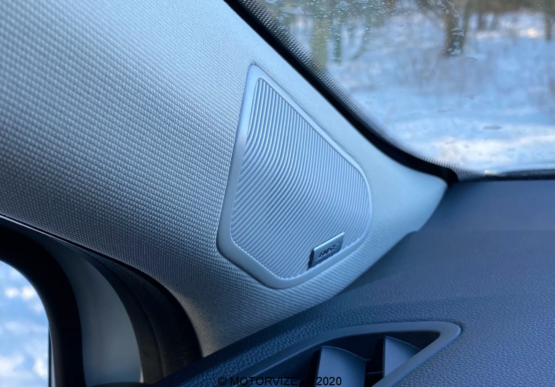 A 2020-as Renault Captur belső terének közeli felvétele, amely a jobb első ajtó hangszóróját hangsúlyozza texturált, hálószerű rácsozattal, körülötte kontrasztos anyagokkal és márkaemblémával, a jármű belsejéből készítve.