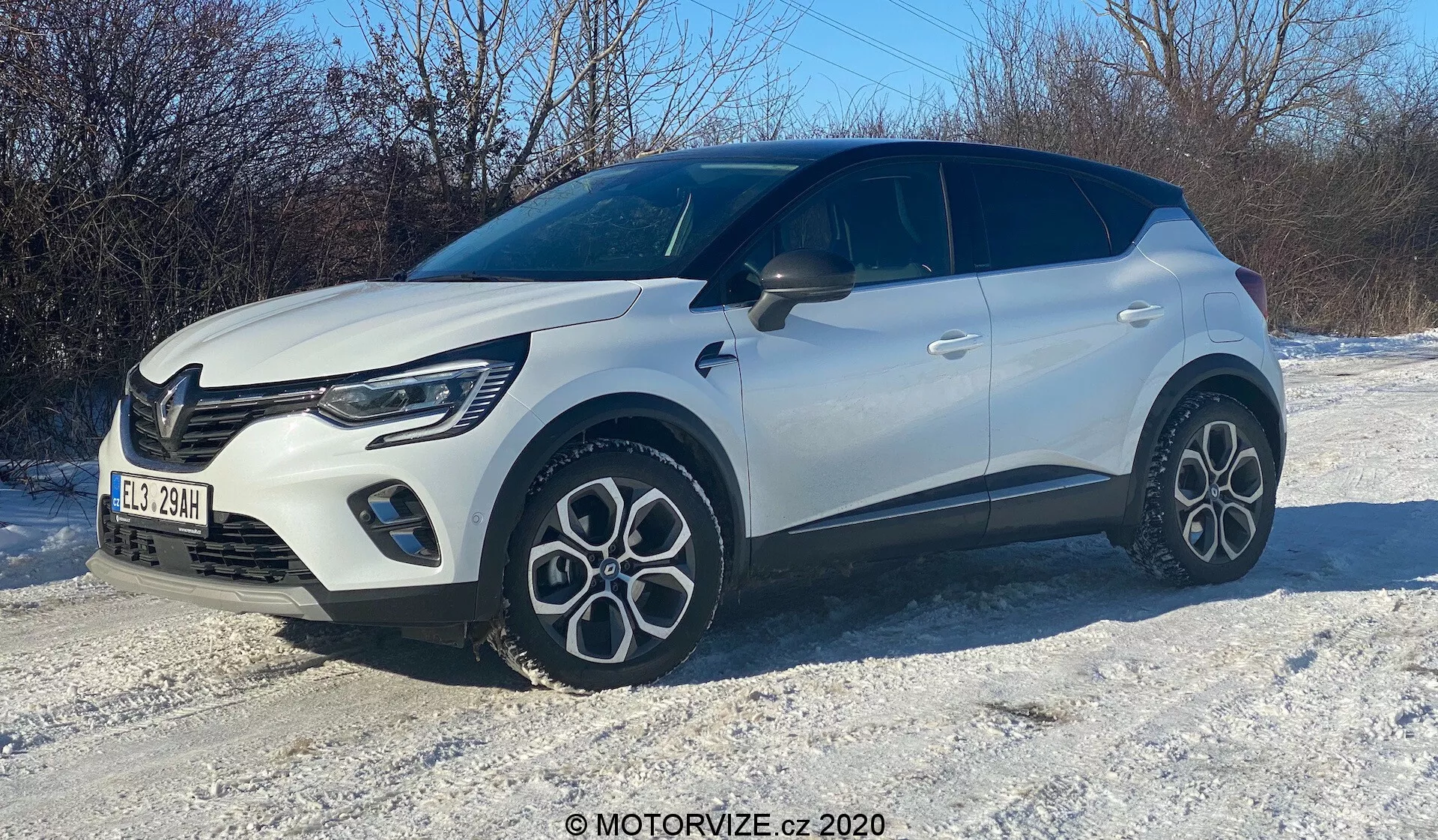 Egy fehér kompakt SUV képe havas terepen, háromnegyedes elülső szögből fényképezve. Látható a jobb oldal, a motorháztető, a hűtőrács, az elegáns LED fényszórók, az első kerék és sárvédő, az utas ajtaja és a hátsó rész egy része. A jármű modern dizájnnal, merész vonalakkal, jellegzetes, többszálas könnyűfém kerekekkel, két tónusú karosszériával, fekete tetővel és jellegzetes hűtőrács mintával rendelkezik, látható nappali menetfényekkel.