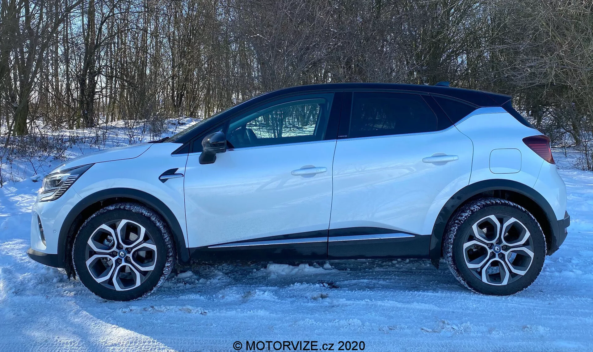 Oldalnézet egy 2020-as Renault Captur-ről, amely jeges vagy havas felületen parkol. Az autó profilja könnyűfém kerekeket, szabványos közúti gumiabroncsokat, négy ajtót, oldalsó ablakokat, köztük egy kis hátsó negyed ablakot és egy irányjelzővel integrált oldalsó visszapillantó tükröt mutat. A tetővonal elöl magasabban kezdődik és kissé hátrafelé lejt, ami egy szabadidőautó vagy ferdehátú kialakítást javasol. A jól megvilágított jelenet, valószínűleg természetes napfény miatt, meghatározza az autó alakját a vidéki vagy külvárosi környezet fáival a háttérben. A kép 'MOTORVIZE.cz 2020' vízjelet tartalmaz.