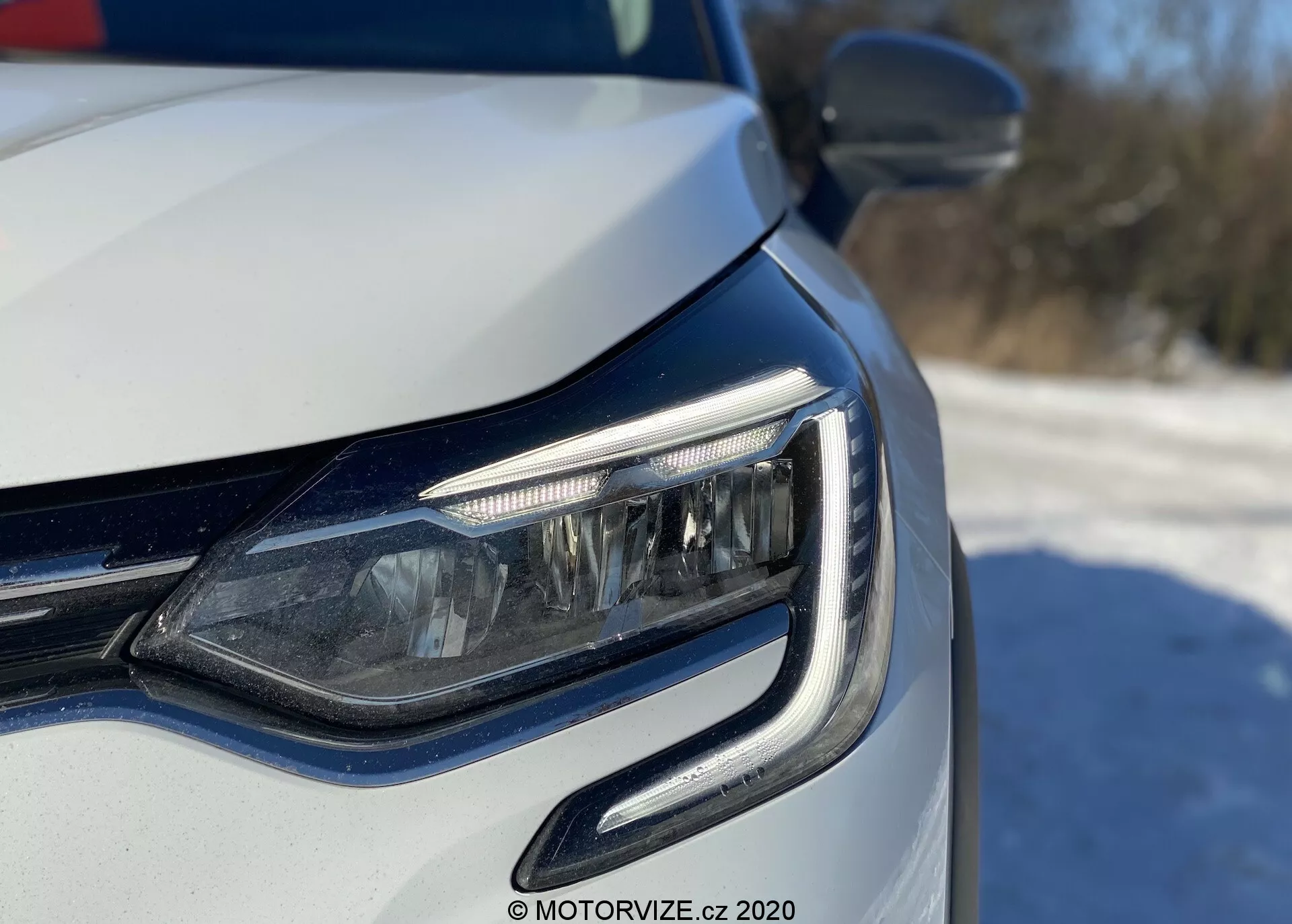 Egy modern autó elülső bal sarkának közeli nézete, amely az LED fényszóróra és a hűtőrácsra fókuszál, alacsony szögből, a lámpa szintje közelében készült. A nappali menetfények ívelt kialakításúak, amelyek körülölelik a fényszóró egységet. Láthatók adaptív fényszórók vagy kanyarvilágító funkciók, fémes festékbevonattal és az integrált irányjelzőkkel a külső visszapillantó tükörben. Az erős természetes fény nappali kültéri környezetet sugall.