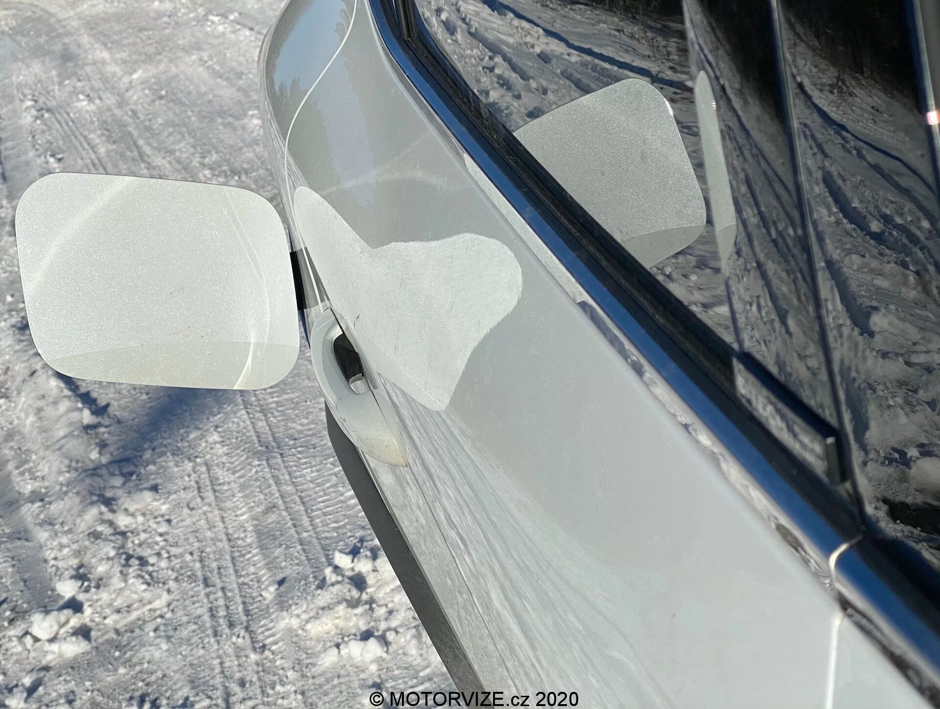 Közelkép egy 2020-as Renault Captur oldalnézetéről havas kültéri környezetben. A fókusz a jobb oldali visszapillantó tükörre irányul, amely előre van hajlítva, ami automatikus vagy manuális hajtogatási funkcióra utalhat. A tükör burkolatán két átlátszó, párhuzamos formájú védőfólia található, valószínűleg szállítás vagy mosás közbeni védelem céljából. Az autó sötét, fényes árnyalatú, például fekete vagy sötétkék, jól karbantartott állapotban van, a tükröződések tiszta, fényes felületet sugallnak. A környező föld és az autó oldala hóval van borítva. Látható az első ajtókilincs egy része is, melyen kulcs nélküli bejutásra szolgáló gomb vagy érintésérzékelő található.