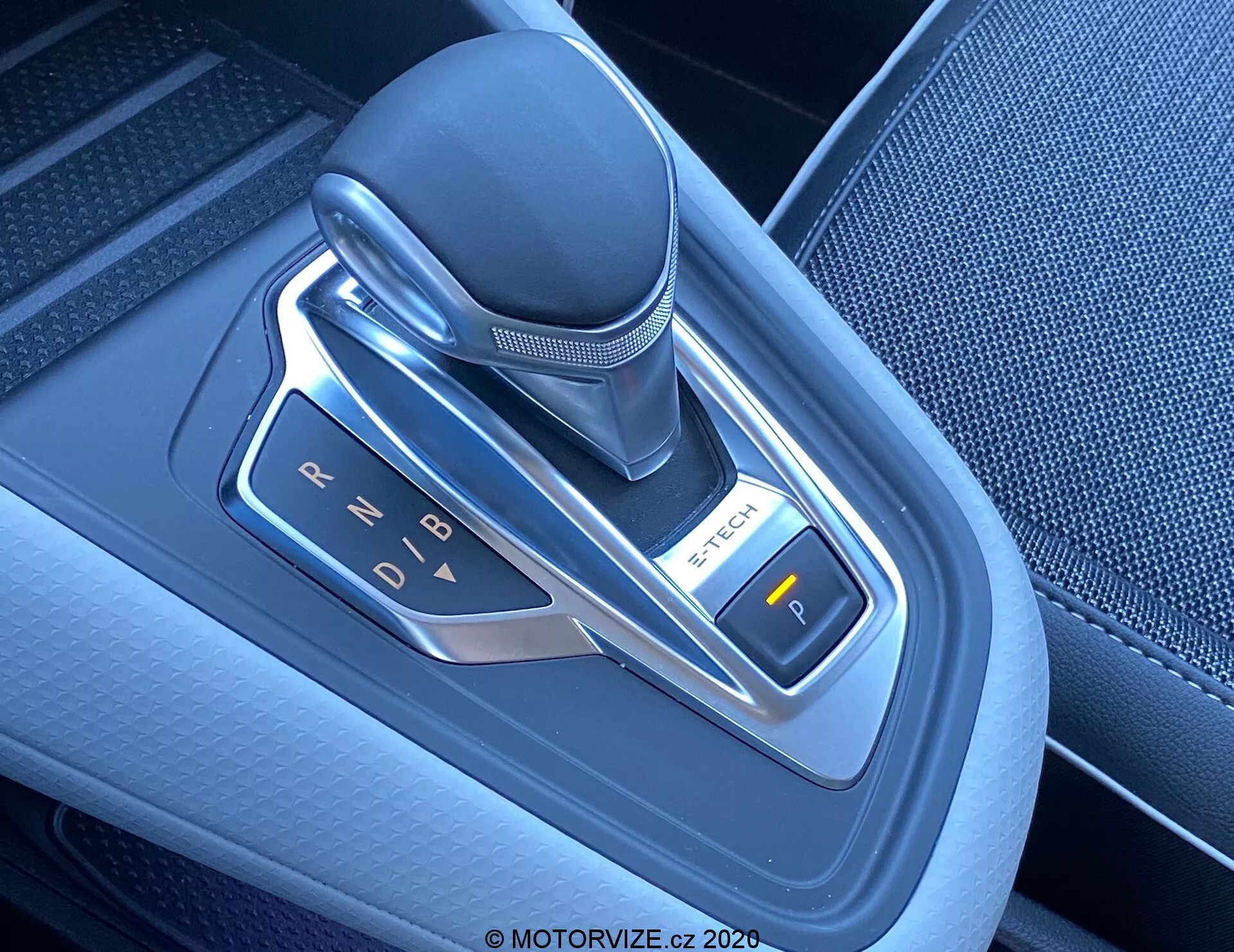 Közelről készült kép egy modern automata sebességváltó karról egy 2020-as Renault Captur-ben, amely a középkonzolon található. Az elektronikus sebességváltó függőlegesen helyezkedik el, és bőrből, műanyagból és fémes díszítésekből készült. A felvétel kissé jobbra dőlő felülnézetből készült, kiemelve a sebességfokozat jelzéseket (P, R, N, D, B), amelyeket LED fények világítanak meg. A jobb oldalon egy 'P' feliratú gomb látható, amely valószínűleg az elektronikus rögzítőfékhez tartozik. A sebességváltó körüli terület magas minőségű texturált műanyagból készült fémes díszítésekkel, amelyek az autó modern technológiáját és belső terének korszerű kialakítását tükrözik.