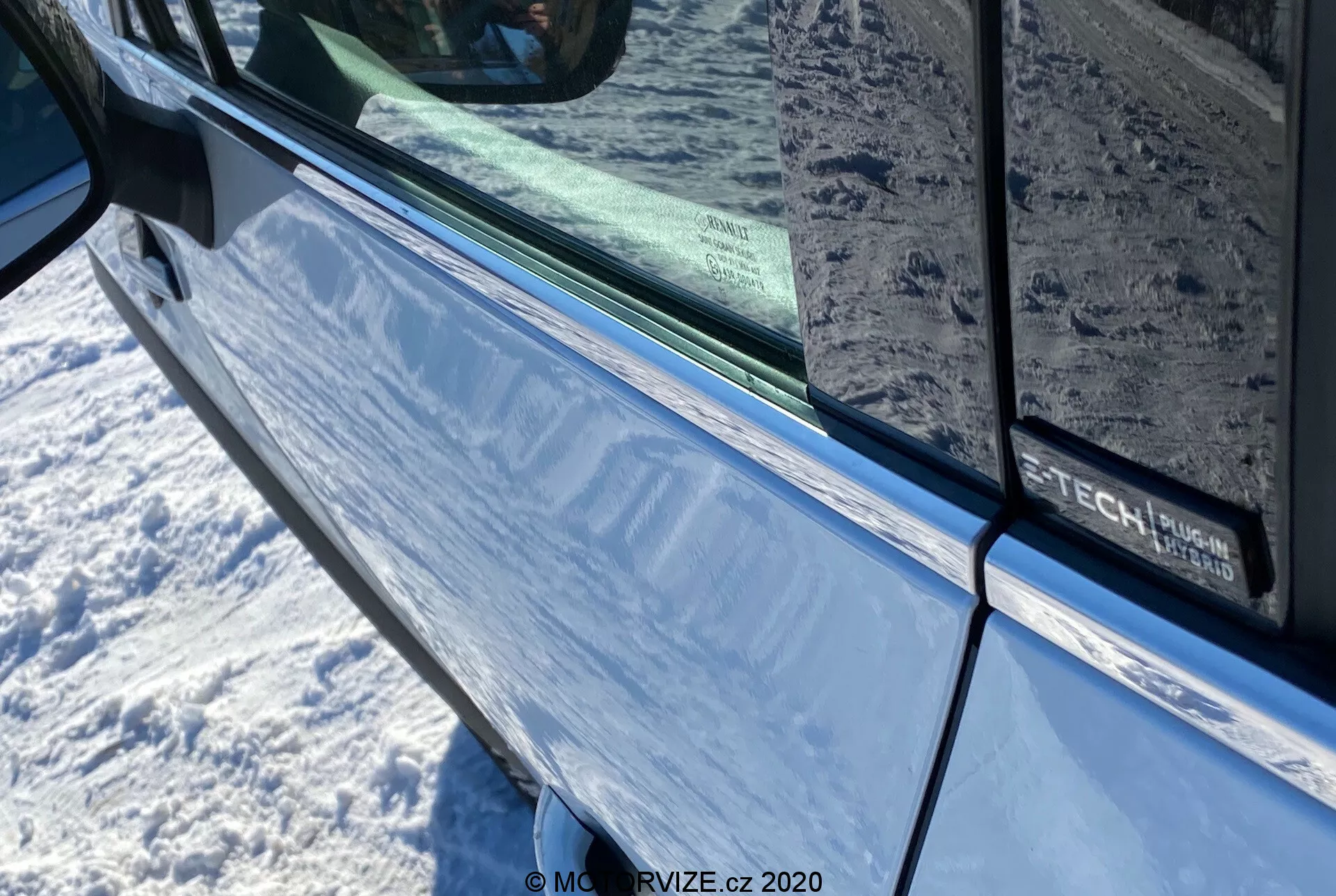 Közelkép a 2020-as Renault Captur-ról fémes kék színben, a vezető ajtajára és az integrált irányjelzővel ellátott oldalsó tükörre fókuszálva. A kép kiemeli az ajtó területét az árnyékolt ablak széléig futó fekete szegéllyel, mely tükrözi a környező környezetet. Az ajtón részben látható egy felirat, amely valamilyen felszereltségi csomagra vagy technológiai csomagra utalhat.