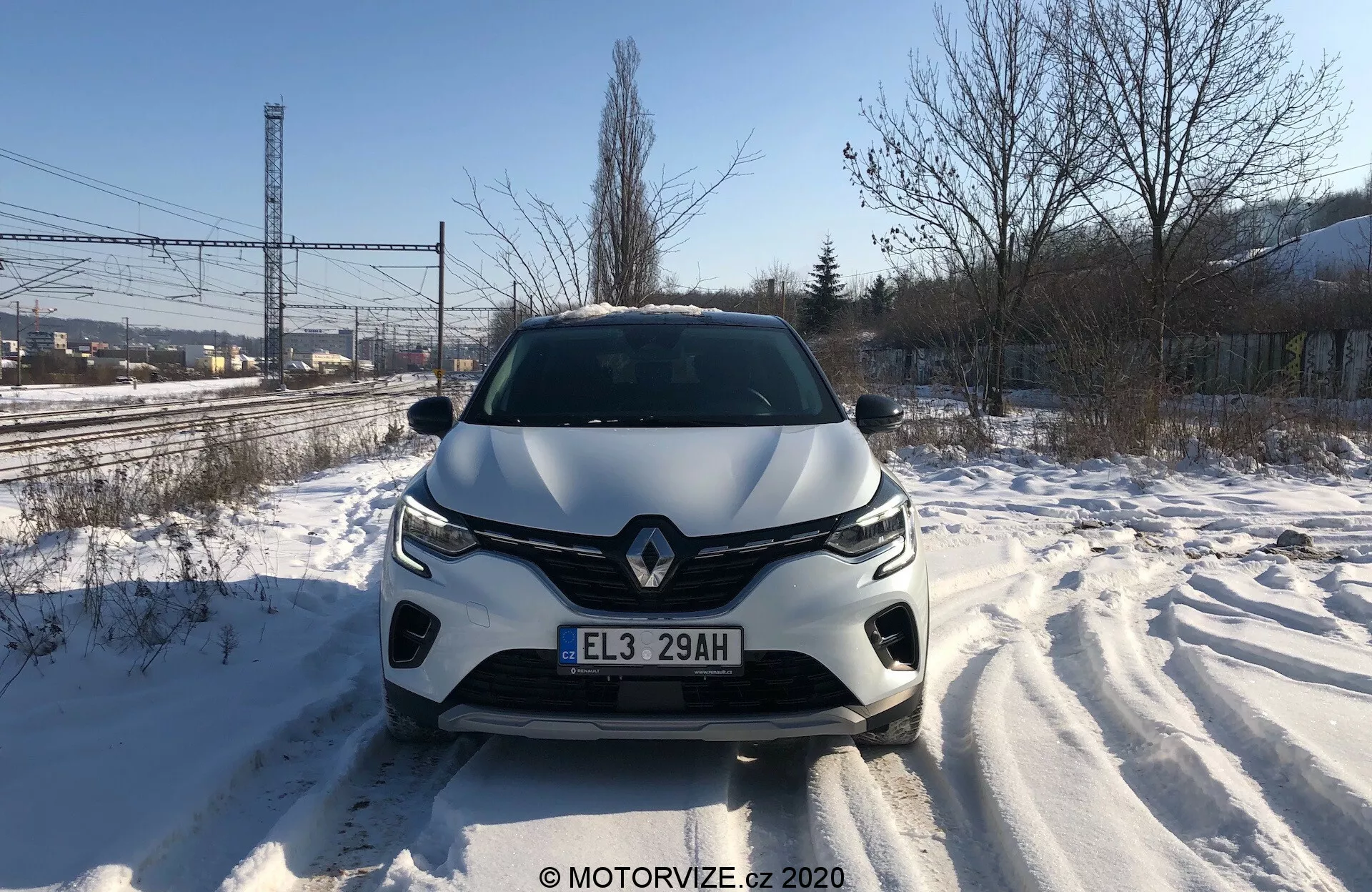 Egyenesen szemből készült fotó a Renault Captur 2020-as modelljéről egy hóval borított úton, nappali megvilágításban, amely részletesen megmutatja az első hűtőrácsot, fényszórókat, formázott motorháztetőt és első lökhárítót. A látható keréknyomok nemrégiben történt mozgást jeleznek. A jármű dizájnja aerodinamikus stílust sugall, kiemelkedő márkalogóval és fejlett világítási rendszerrel. Az első lökhárítóba vezetéstámogató szenzorok vagy kamerák is beépítve lehetnek.