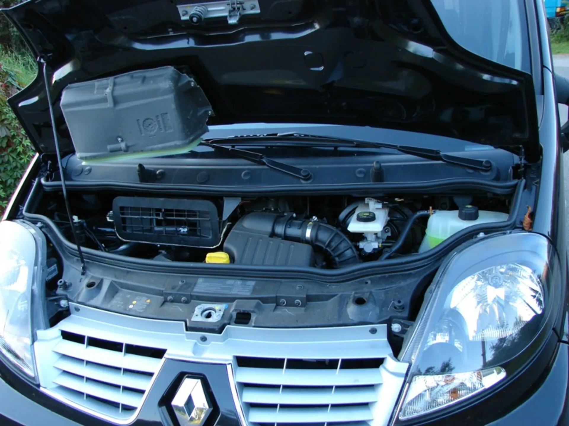 2006-os Renault Trafic Combi első háromnegyedes nézete nyitott motorháztetővel, amely felfedi a motoros részt és az alkatrészeket, beleértve a motorblokkot fedéllel, légszívót, és folyadéktartályokat, hagyományos halogén fényszórókkal szegélyezve