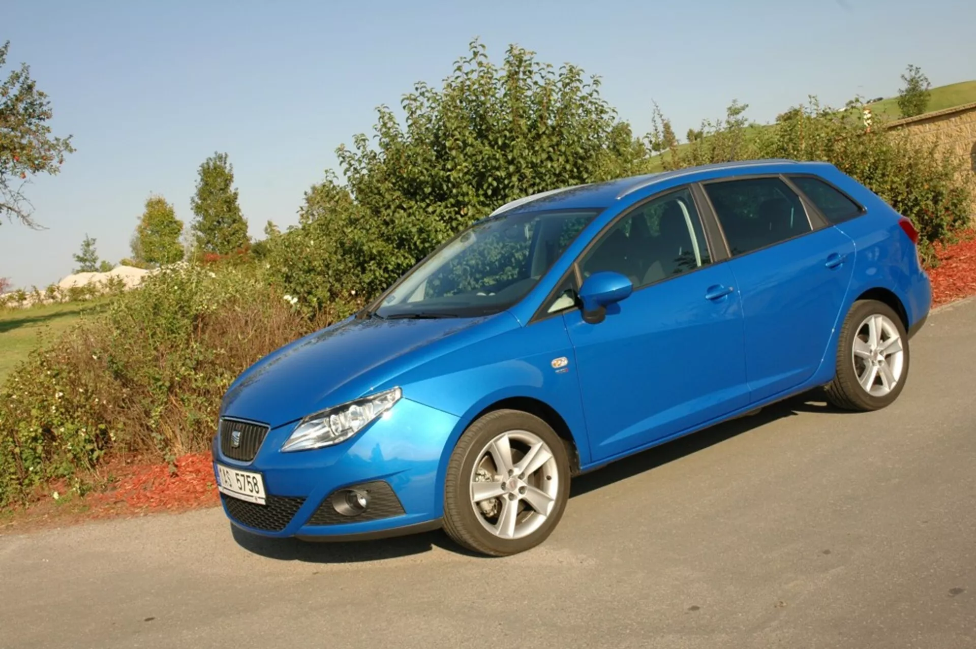 Világoskék Seat Ibiza 2012 ferdehátú parkolva az aszfalton a zöldellő háttér előtt, bemutatva aerodinamikus első hűtőrácsát, hátrafelé kunkorodó fényszóróit és oldalsó profilját többszólemezes ötvözött kerekekkel a természetes nappali fény alatt