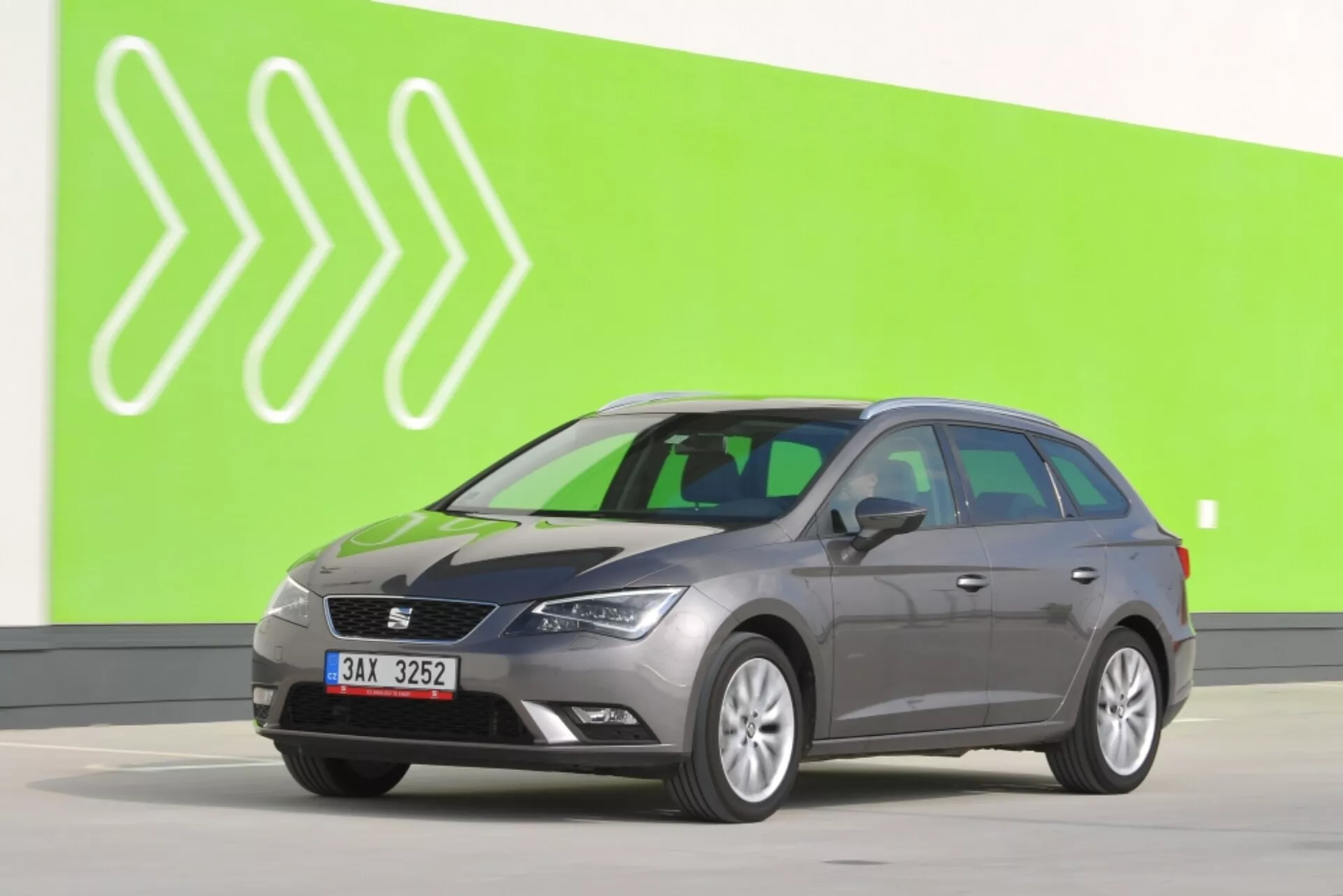 Szürke Seat Leon SC 2012 kombi teljes oldalnézetben zöld hátteren stilizált fehér nyílhegyekkel, oldalsó tükrökkel forduló jelzőkkel, jellegzetes ajtókontúrokkal és standard keréktárcsákkal világosszürke stúdiópadlón