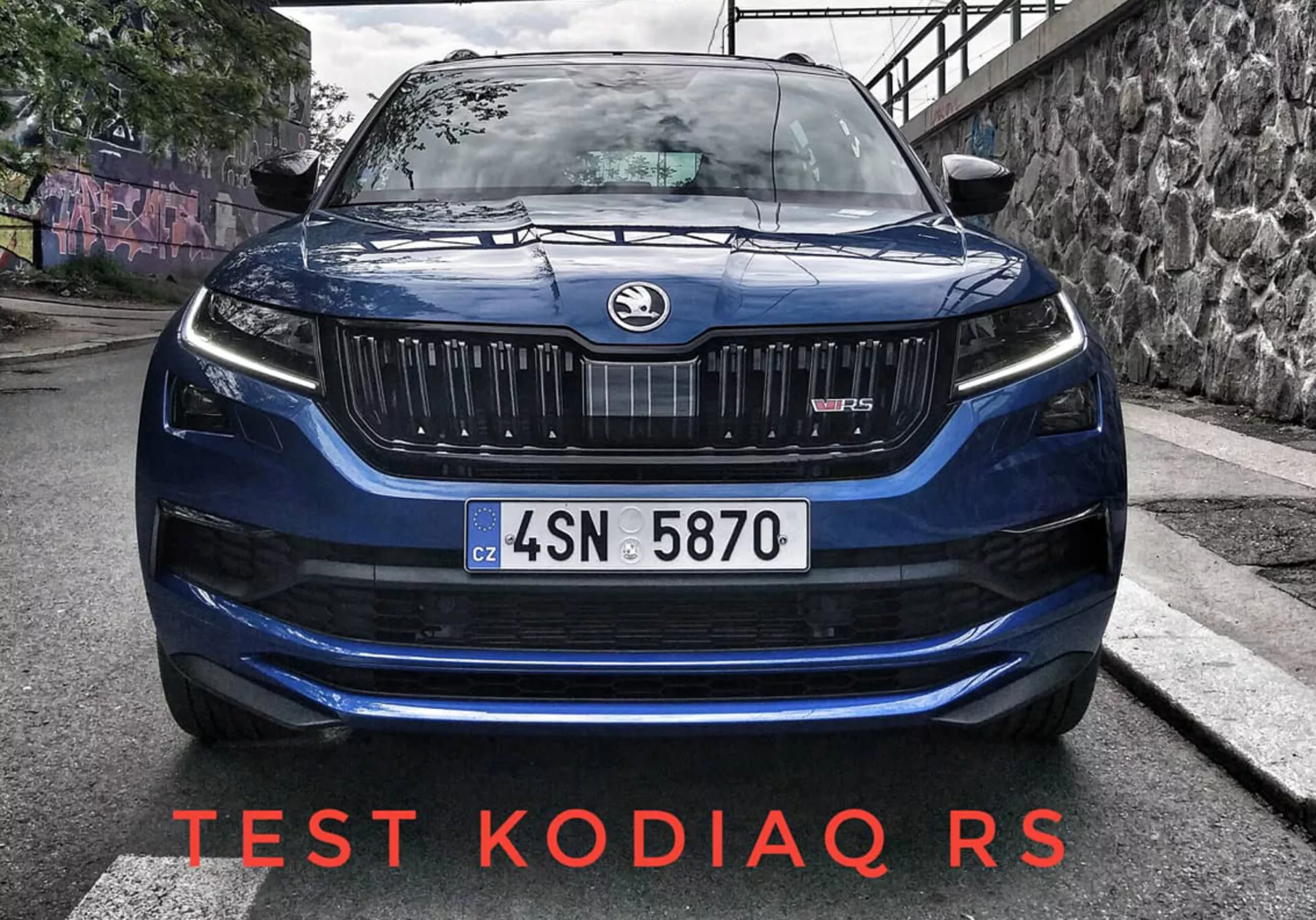 Közeli első nézet egy 2017-es Skoda Kodiaq Scout-ról, amelyet domináns rács jellemzi függőleges lamellákkal, elegáns LED fényszórók, finoman domború motorháztető a jobb aerodinamika érdekében, és hatékony légbeömlők a lökhárítón, városi úton graffiti háttérrel természetes nappali fényben