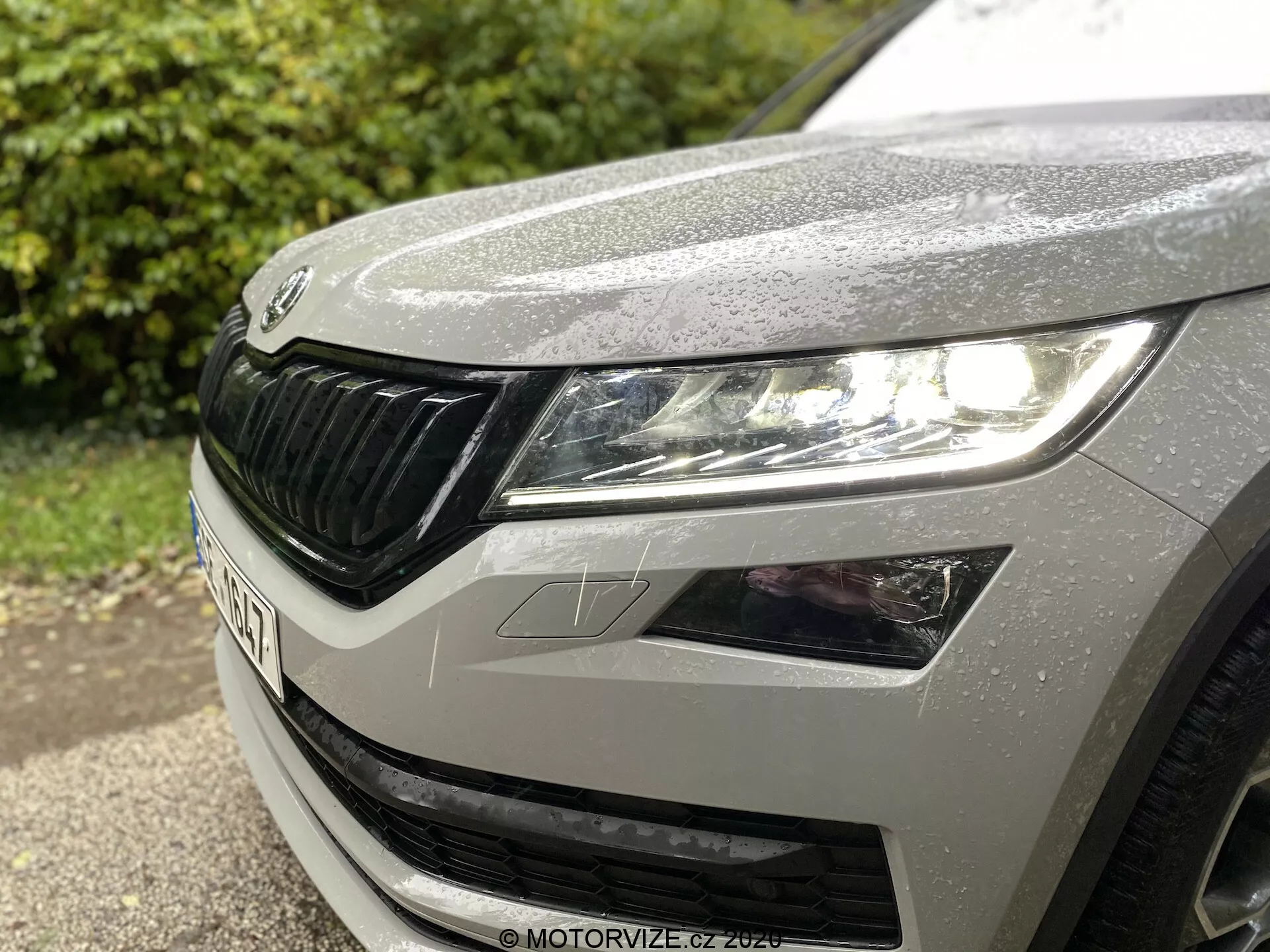 Közelkép egy 2017-es Škoda Kodiaq bal elülső sarkáról, a modern fényszóróegységgel, LED-technológiával és integrált nappali menetfényekkel, a jellegzetes rács függőleges lamelláival és a ködlámpa részletével. A fémes ezüst vagy szürke festék vízcseppekkel díszített, kiemelve az elegáns, aerodinamikus formatervezést egy alacsony szögű, átlós perspektívából.