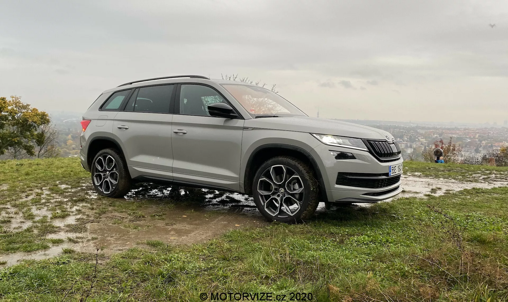 Ezüstszínű 2017-es Skoda Kodiaq SUV füves, kissé sáros talajon parkol, oldal-bemutató szögből rögzítve. Tiszta kilátás az első rácsra, fényszórókra, bal oldali profilra, elegáns fényszóró dizájn LED technológiával, jellegzetes rácsdizájn, a rács alatti lehetséges szenzorok asszisztencia rendszerekhez, oldal tükrök irányjelzőkkel és esetleges holttér-figyeléssel, nagyméretű többágú alufelnik, az utakra és mérsékelten terepre is alkalmas gumiabroncsokkal, amelyek összkerékhajtási képességeket javasolnak. Egy lejtőn helyezkedik el, távoli, enyhén ködös városképpel a háttérben borús ég alatt, a puha világítás azt sugallja, hogy korai reggeli vagy késő délutáni időpontban készült. Az emberek a háttérben élvezik a kilátást, kiemelve a szabadidős és életstílus-fókuszú vonzerőt.