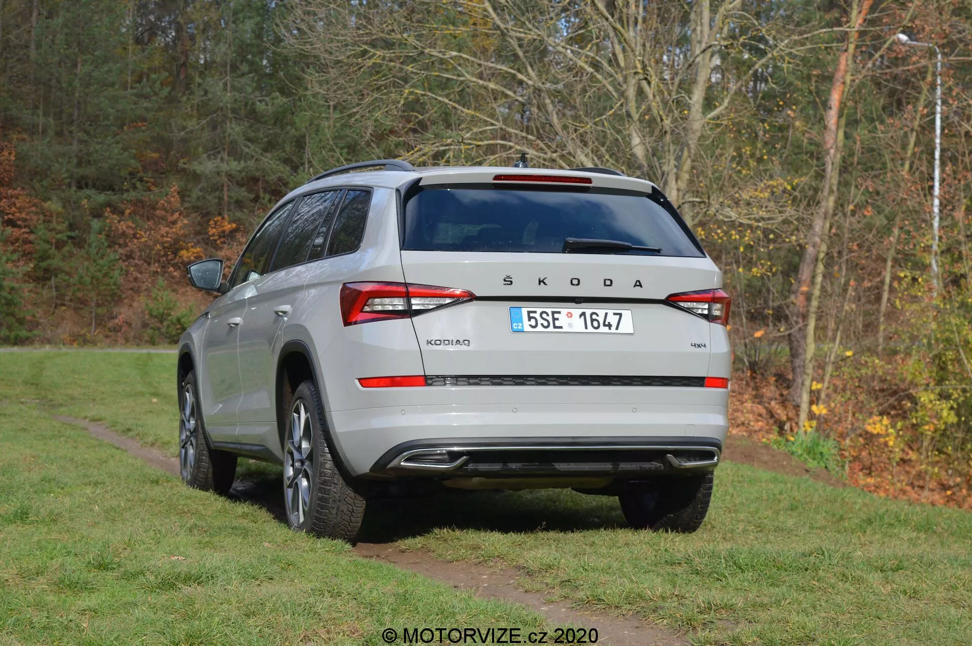 Háromnegyed hátulnézet egy ezüst Škoda Kodiaq 2017-es SUV-ról, ami egy füves területen parkol, fák hátterével. A jármű tetőre szerelt légterelővel, cápauszony antennával, hátsó ablaktörlővel és ablakfűtővel rendelkezik. A hátsó ajtón jól látható a Škoda embléma és a 'Kodiaq' modellnév, európai stílusú rendszámtáblával. A körbefutó hátsó lámpák LED-technológiát sugallnak. A lökhárító parkolószenzorokat, fényvisszaverőket és hátsó védőlemezt tartalmaz. Láthatóak a kettős kipufogórendszer körvonalai, és az oldalsó profilon ötvözet kerekek és védőburkolatok találhatók.