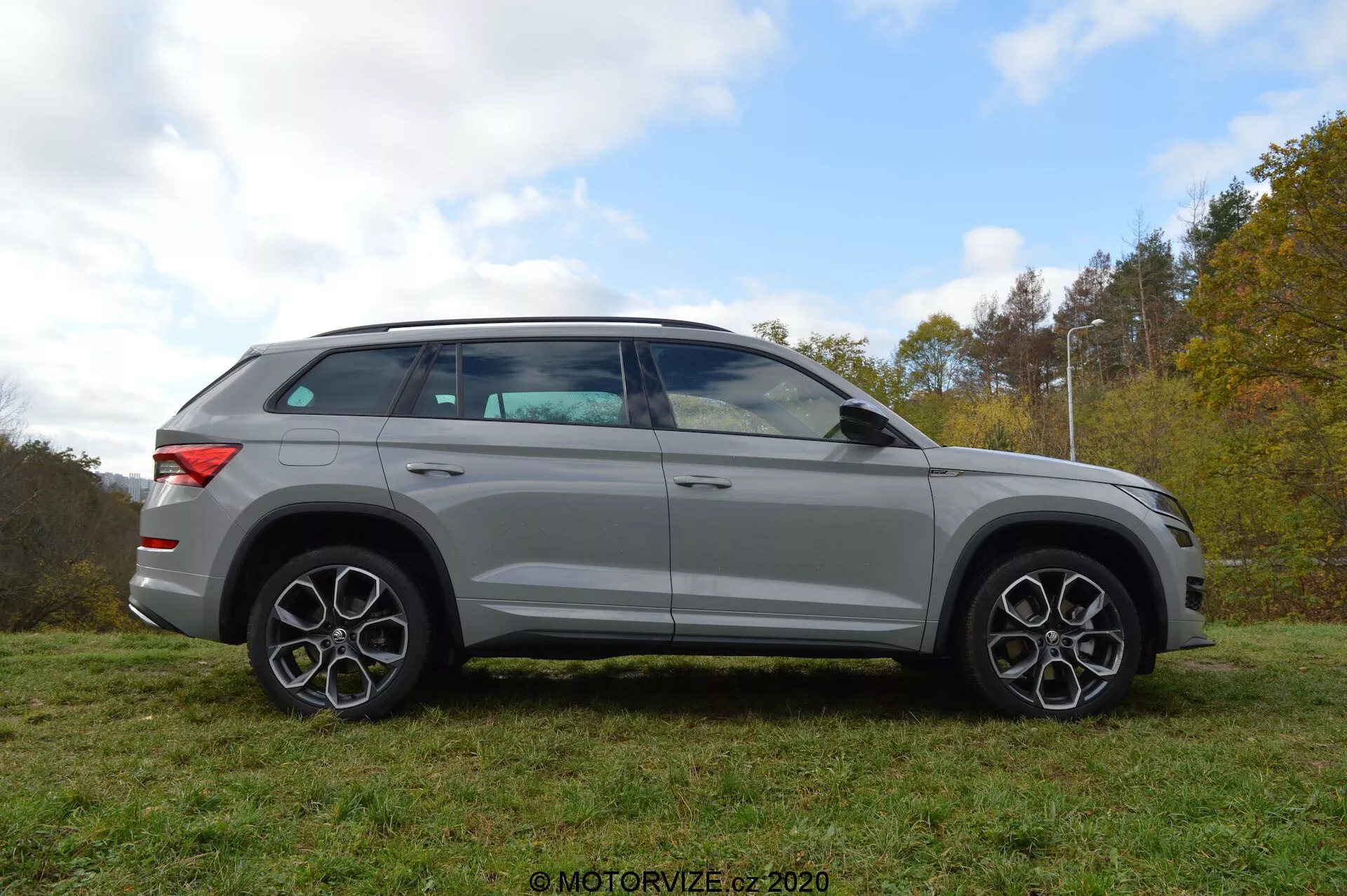 Egy ezüstszínű Skoda Kodiaq 2017 teljes oldalsó profilképe, amely egy füves területen parkol, háttérben fákkal és felhős éggel. A jármű dizájnja merész vonalakat, nagy könnyűfém kerekeket és jellegzetes többlapkás felniket tartalmaz. A tükrök, melyek integrált irányjelzőkkel rendelkeznek, közvetlenül az ablakvonal alatt vannak rögzítve. Az ablakok enyhén sötétítettek, és a tetővonal lágyan lejt hátrafelé, ami sportos, kupészerű profilt mutat. Az SUV tetősínrendszerrel is felszerelt.