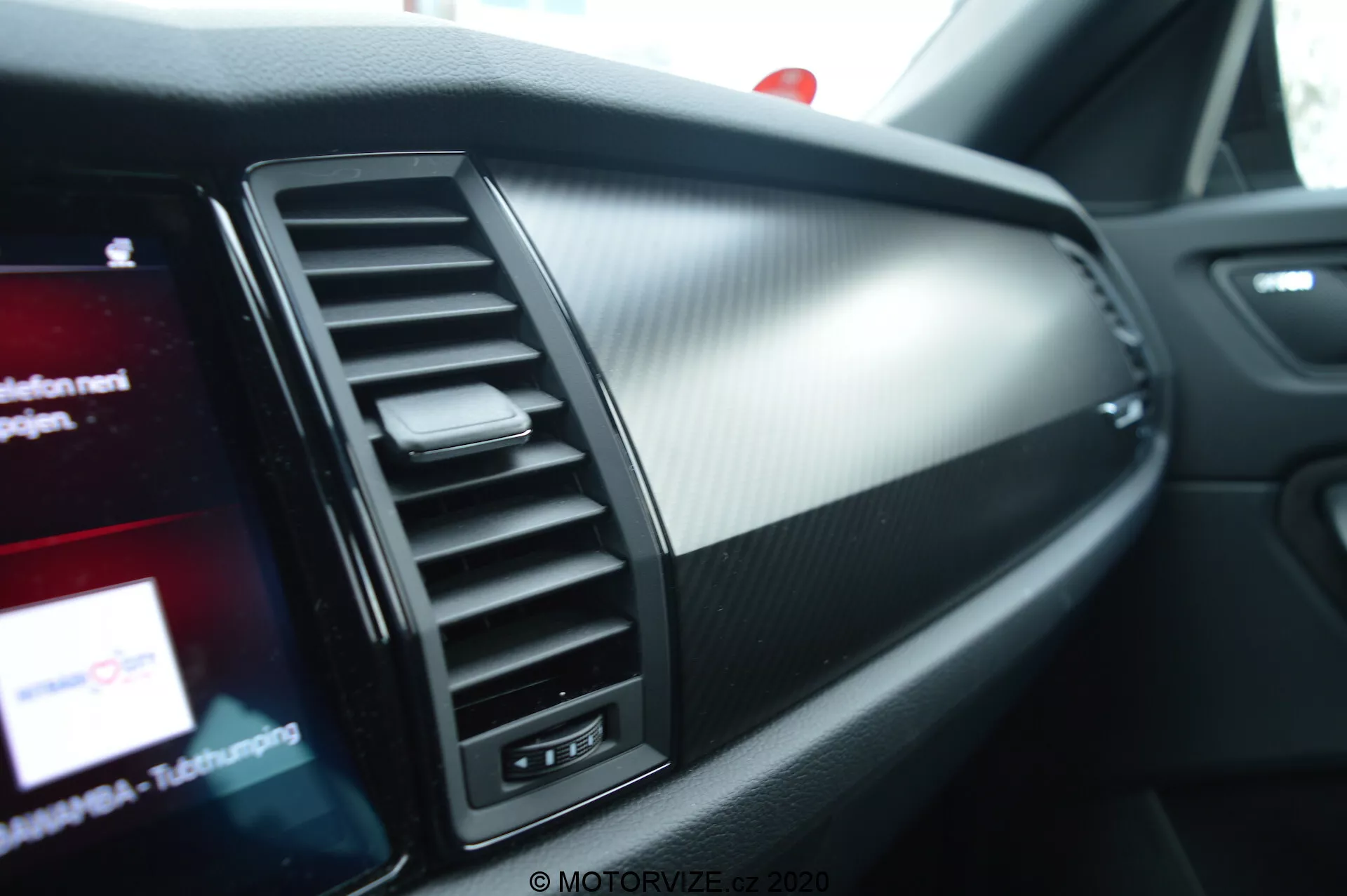 2017-es Skoda Kodiaq belső részlet: az utas szemszögéből nézve, az első utas oldali műszerfal területére összpontosítva, légkondicionáló szellőzővel, csiszolt fém díszítéssel, karbon mintázatú textúrával, részben látható infotainment képernyővel, utasoldali légzsák jelzővel és hangulatvilágítással.