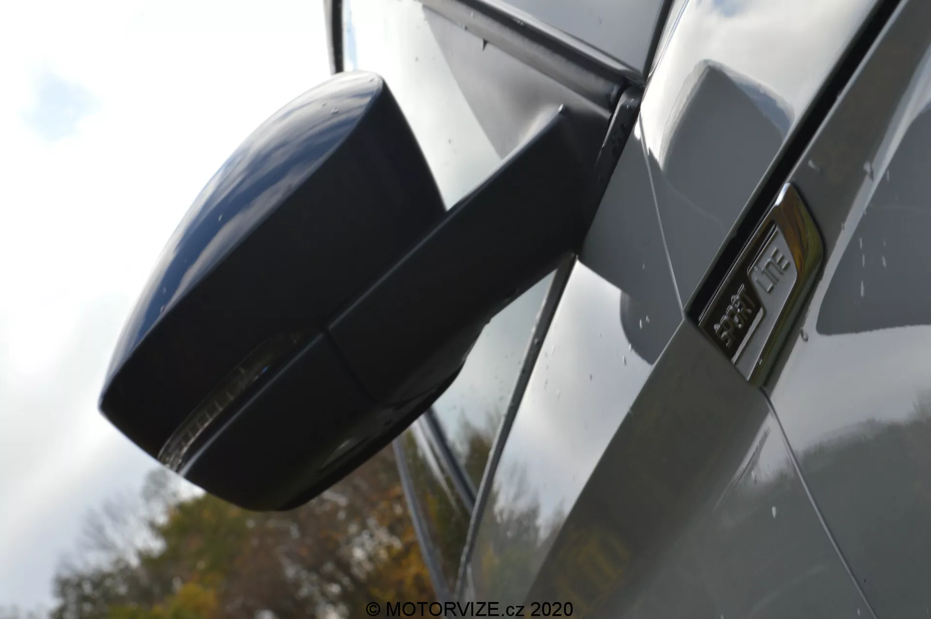 Közelkép egy 2017-es Skoda Kodiaq oldalsó visszapillantó tükréről és vezető ajtajának területéről, kiemelve a modern, aerodinamikus kialakítást fényes festett felületekkel és esetleg fekete műanyag vagy karbon szálas textúrával, kiemelve a tükröt és egy emblémát az ajtón. A háttér defókuszált, növelve a tükör részleteit borongós megvilágításban.