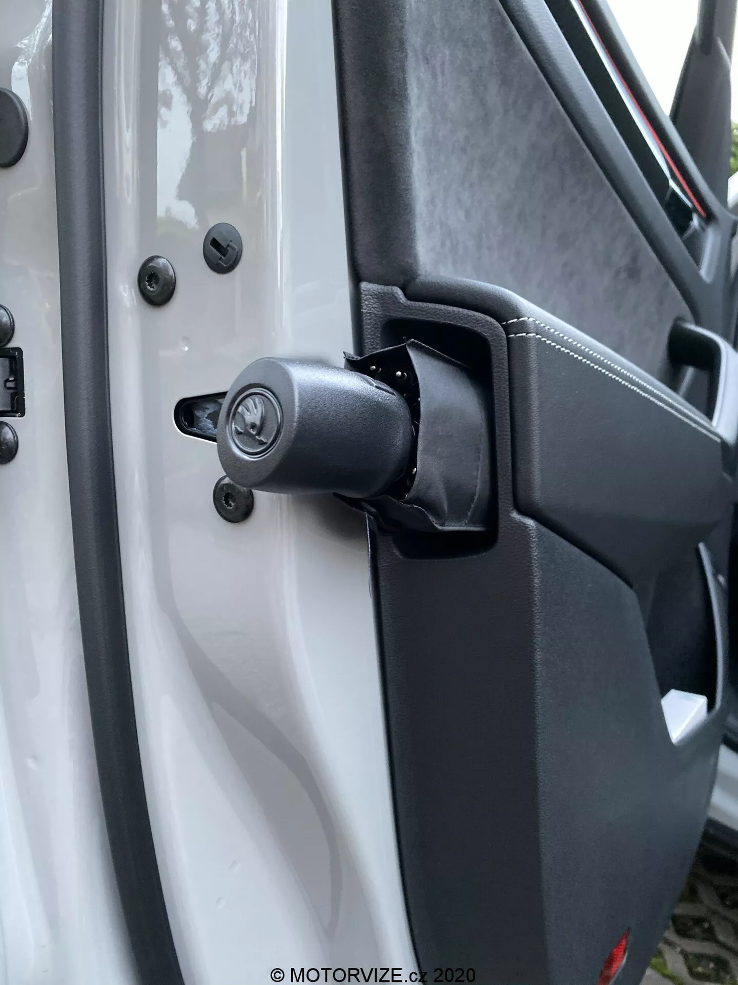 Közelkép a 2017-es Škoda Kodiaq ajtózsanérjának robusztus mechanizmusáról és a környező területről, amelyen látható egy rugós elem, jelezve a nagy teherbírású használatot. A fehér jármű kontrasztot képez a fekete zsanér elemekkel, beleértve a hagyományos kulcslyuk-zár mechanizmust a zsanér felett, amelyet hatszög fejű csavarokkal rögzítettek a tartósság érdekében. Az ajtó belső panelének texturált műanyag borítása és a biztonságos rögzítési pontok fokozzák a praktikus dizájnt. Az elmosódott háttérelemek kiemelik a zsanért és a zármechanizmust.