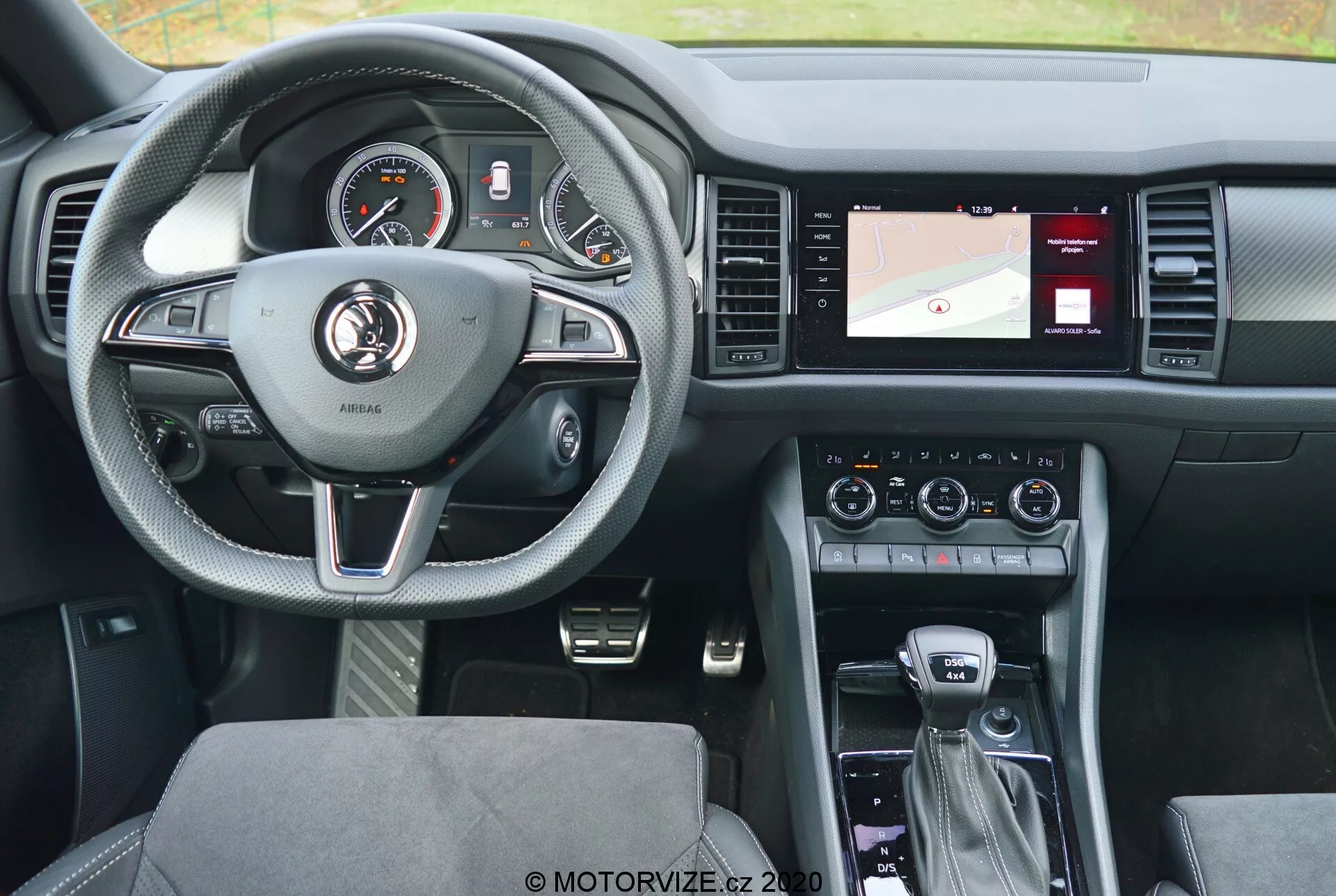 Egy 2017-es Skoda Kodiaq belső nézete a hátsó ülésből, kiemelve a bőrborítású háromküllős kormánykereket ezüst díszítésekkel és a sebességtartó automatika, az audió és a kihangosító kommunikáció gombjaival. A kormány mögött található a műszercsoport, amely analóg számlapokat és digitális kijelzőt tartalmaz. A jobb oldalon található középkonzol navigációs térképet, klímavezérlést és hangbeállításokat tartalmaz. A sebességváltó kar automata váltót jelez, további vezérlőkkel az infotainment rendszerhez és a vezetési üzemmódokhoz. A modern kialakítás célja a kényelem és a technológia egyensúlyának megteremtése, mintás szövet ülésekkel és jól megvilágított, felhasználóbarát elrendezéssel.