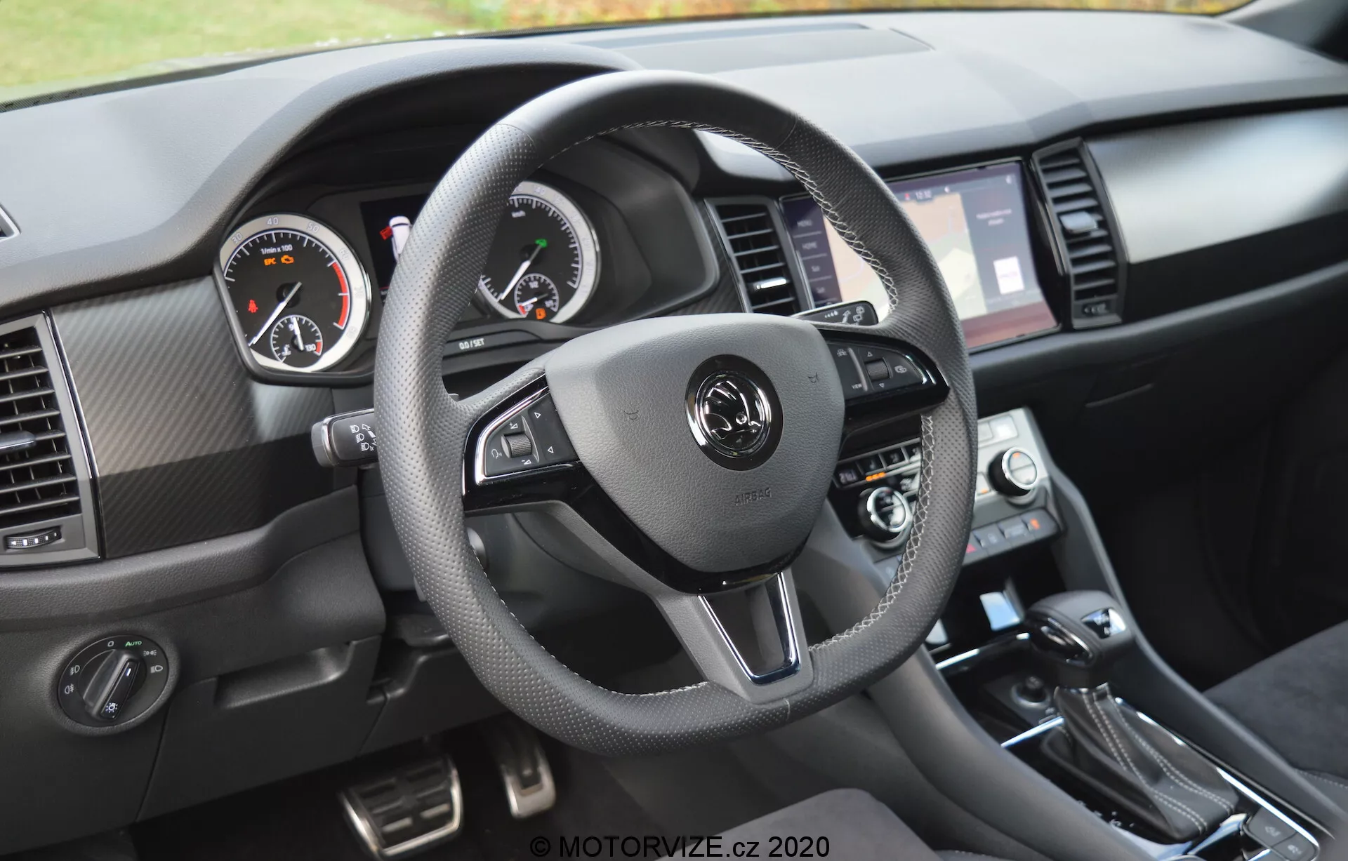 Egy 2017-es Skoda Kodiaq belső nézete a műszerfal vezetői perspektívájából. A háromküllős kormánykerék, amely bőrre emlékeztető anyaggal van bevonva kontrasztos varrással, beépített vezérlőkkel rendelkezik audio, kihangosítás és tempomat beállításokhoz. Az analóg műszerek, bal oldalon a sebességmérővel és jobb oldalon a fordulatszámmérővel, egy középen elhelyezett digitális kijelzővel egészülnek ki. Az információs és szórakoztató érintőképernyő, amelyet szellőzőnyílások közrefognak, a klímavezérlő kapcsolók és a váltókar jól láthatóak, valamint texturált anyagok és csiszolt fémes díszítések.