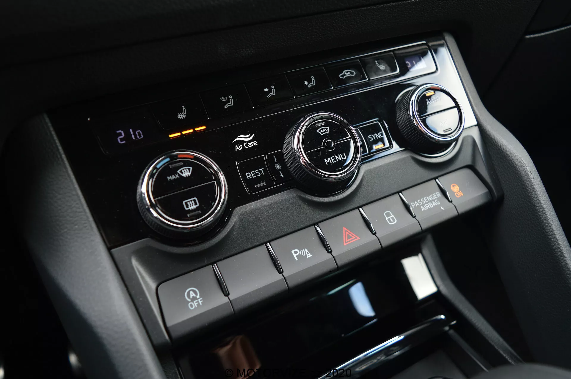 Közeli nézet a 2017-es Škoda Kodiaq középkonzoljáról, kiemelve a klímavezérlő rendszert két nagy hőmérsékletszabályzó gombbal és egy digitális kijelzővel, amely 21 Celsius fokra van állítva. A képen láthatók továbbá légkondicionáló, levegőkeringtető, ablakpárátlanító gombok és egy 'MENU' gomb. Alatta billenőkapcsolók találhatók parkolószenzorok, automatikus start/stop letiltás és vészvillogók számára.