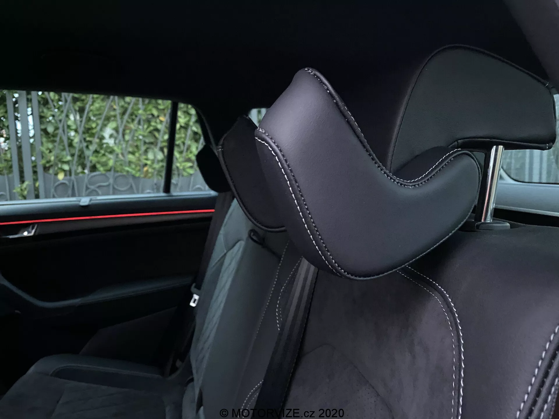 A 2017-es Skoda Kodiaq belső nézete, amely egy autóülésre összpontosít. A szögletes felvétel kiemeli a fejtámla részét, amely fekete anyaggal, valószínűleg bőrrel vagy kiváló minőségű szintetikus anyaggal van kárpitozva, markáns kontúrral a kényelem érdekében. A pontos varratok kontrasztos cérnával luxus érzetet keltenek. A háttérben vízszintes vörös környezeti megvilágítás látható, ami kiemeli a modern autóipari design elemeit. A látható hátsó utastér-kényelmi elemek közé tartozik a biztonsági övek és esetleg egy középső kartámasz, ami a második üléssorra utal.