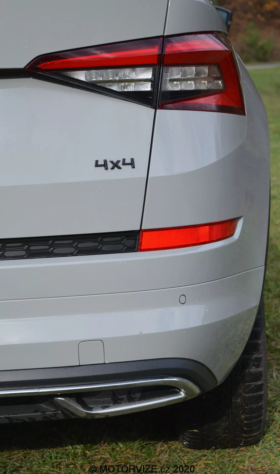Közelképek egy 2017-es Skoda Kodiaq hátsó sarkáról, a hátsó világítás, feliratozás és lökhárító részleteire fókuszálva. A kép szöge a jármű jobb oldali hátsó részét emeli ki, vízszintesen elrendezett, éles, szögletes hátsó lámpacsoportot mutatva, amely a test oldalára hajlik. A hátsó lámpa alatt látható a hátsó panel és a lökhárító, amely ezüst vagy világosszürke színű, tompított környezetet tükröződik. A lökhárító fekete műanyag alsó részt méhsejt mintázattal és ezüst vagy krómmal díszített elemmel tartalmaz, amely valószínűleg a parkolási segítőrendszerhez vagy a stílushoz kapcsolódik. A hátsó lámpa fölött található '4X4' jelvény a négykerék-meghajtást jelzi. A nagy felbontású kép részletes felületi textúrákat mutat, részben látható gumiköpenyvel az alján, fű fölé helyezve. A '© MOTORVIZE.cz 2020' vízjel arra utal, hogy a kép egy cseh autós kiadvány számára készült.