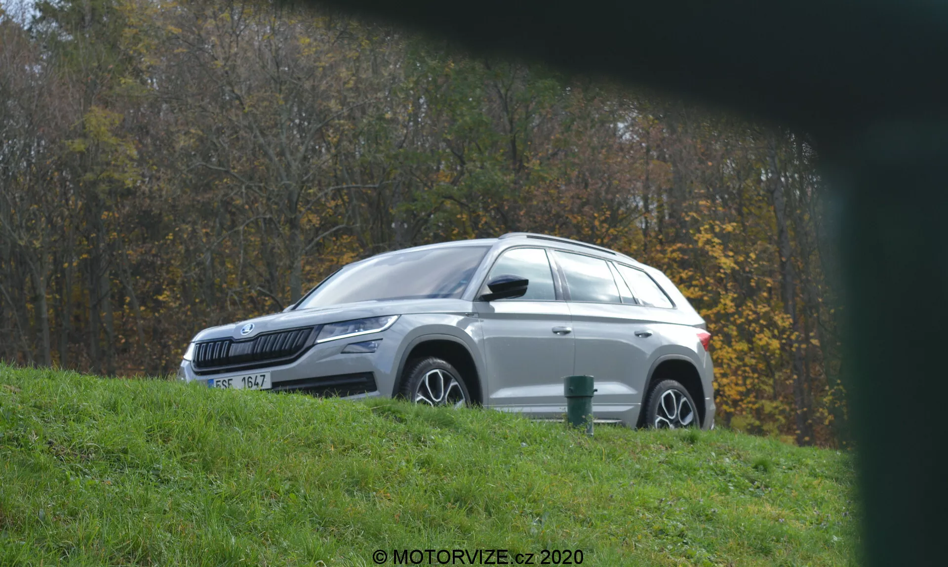 Egy fehér Skoda Kodiaq 2017 SUV oldalsó profilnézete, amit egy füves dombon parkoltak, balra nézve, könnyűfém felnikkel és a napfényt visszaverő fényes fényezéssel. A környezet lombozatot és fákat tartalmaz, amit vidéki vagy parkban levő helyszínt sejtet. Alacsony szögből, elmosódott előtérű nézet, a jármű modern dizájnelemeit mutatja, mint például LED-es fényszórók, integrált irányjelzőkkel ellátott tükrök, színezett üvegek, tetősínek és cápauszony antennát.