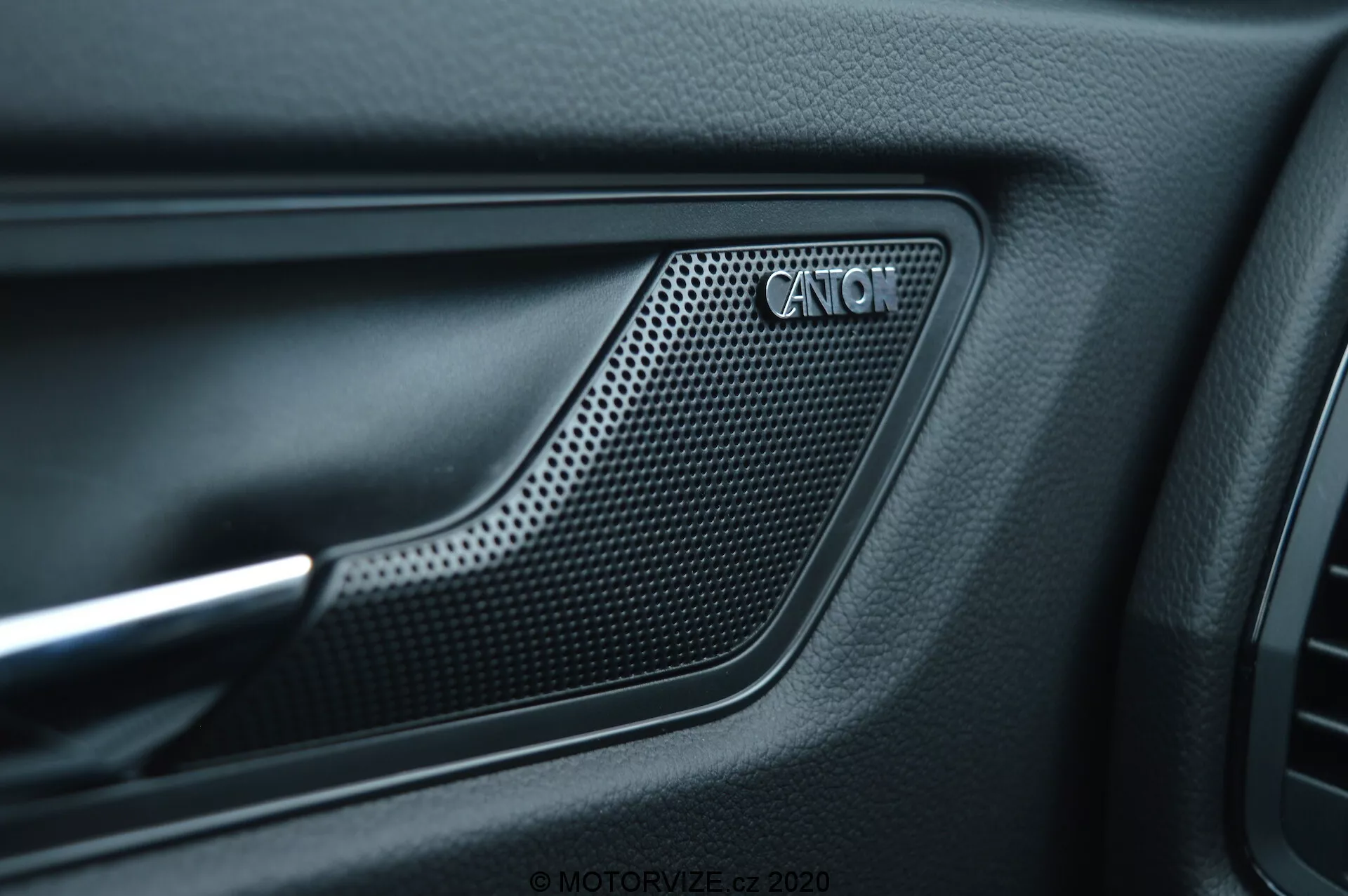 Közelről készült fénykép a 2017-es Skoda Kodiaq belső ajtópanelekével, amelyen a CANTON hangszórórács látható. A rács perforált, méhsejt-szerű mintázatú anyagból készült, és egy benyomott kontúrral keretezett mélyedésben helyezkedik el. A rács körül matt fekete, textúrált anyag található, mellette kontrasztos fém díszítéssel, tükrözve az autó kifinomult belső kialakítását és prémium audiorendszerét.