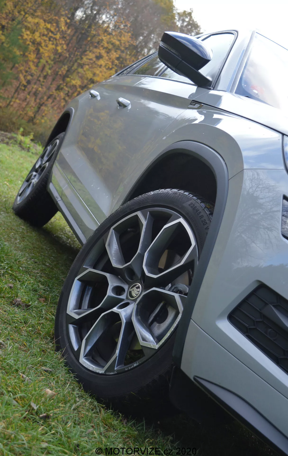 2017-es Škoda Kodiaq közelről, alacsony szögből, az elülső kereket és a sárvédő részt kiemelve. A fűre parkolt autó többágú, könnyűfém kerékkel, integrált irányjelzővel ellátott aerodinamikus oldalsó tükörrel, szögletes karosszériaelemekkel, metál fényezéssel és az első sárvédőn lévő szellőzőnyílással rendelkezik, homályos természetes háttér előtt.