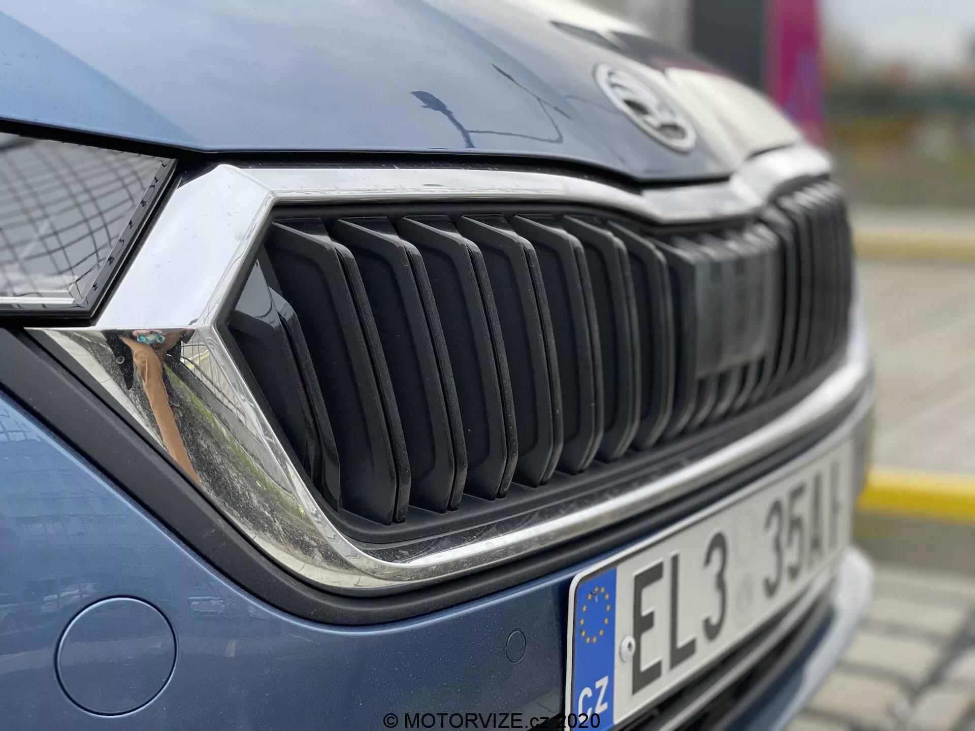 Közeli kép egy autó elülső részéről, amely a hűtőrácsra és a fényszóróra összpontosít. Az kép egy első, jobboldali szögből készült, kissé felfelé nézve, részletesen megmutatva a hűtőrács jobb oldalát, a jobb fényszórót és a motorháztető egy részét. A hűtőrács függőleges lamellái hangsúlyosak, merész dizájnt sugallnak. A bonyolult fényszóró fejlett világítástechnológiát, például LED- vagy adaptív rendszereket sejtet. A motorháztető kissé elmosódott, kiemelve a hűtőrács és a fényszóró textúráját. Az autó jól karbantartott, tükröződésekkel, amelyek felhős, szabadtéri beállítást vagy árnyékot jeleznek.