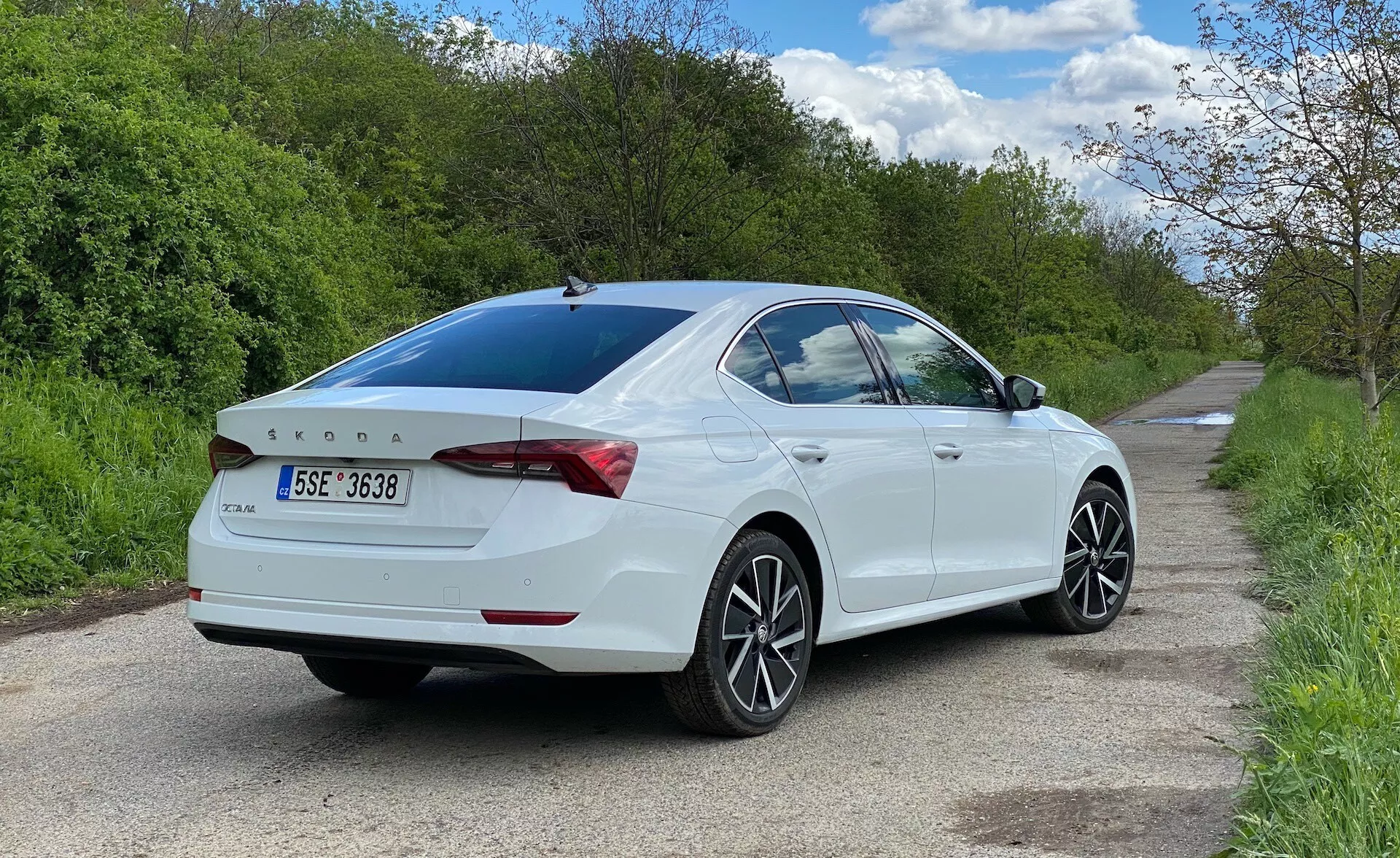 Háromnegyedes hátulnézeti kép egy fehér Skoda Octavia 2020 szedánról, amely egy keskeny út jobb oldalán parkol, és az utasoldali nézetet mutatja alsó szintről. Az autó kitölti a kép nagy részét, kiemelve a dizájnvonalakat, a hátsó lámpát, amely a csomagtartóra kanyarodik, a finoman integrált spoilert, a hátsó feliratot, a többküllős könnyűfém kerekeket, az enyhén sötétített hátsó ablakot és a beépített irányjelzőkkel ellátott oldalsó tükröket.