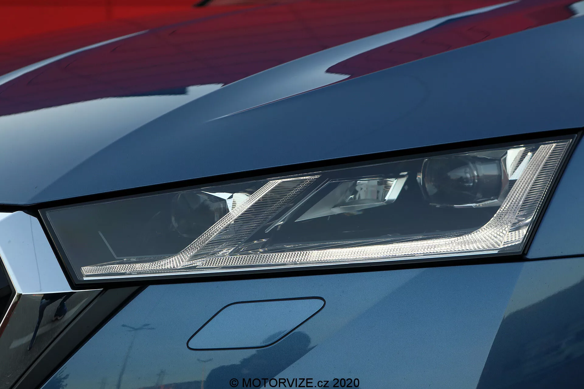 A Škoda Octavia 2020 limuzin modell bal fényszórójának közeli képe, amely kiemeli szögletes dizájnját és a modern LED vagy lézer világítástechnológiát. Az alacsony szögből készült fotó az összetett belső tükröket és a részletes elemeket mutatja a tiszta lencsével borított fényszóróban. A nagy felbontású kép a motorháztető és a sárvédő egy részét is feltárja, amelyen fémes fényezés látható, jelezve a prémium vagy jól karbantartott járművet. Ez a kép a fejlett autóvilágítást és a sportos autókhoz társított agresszív stílust hangsúlyozza.