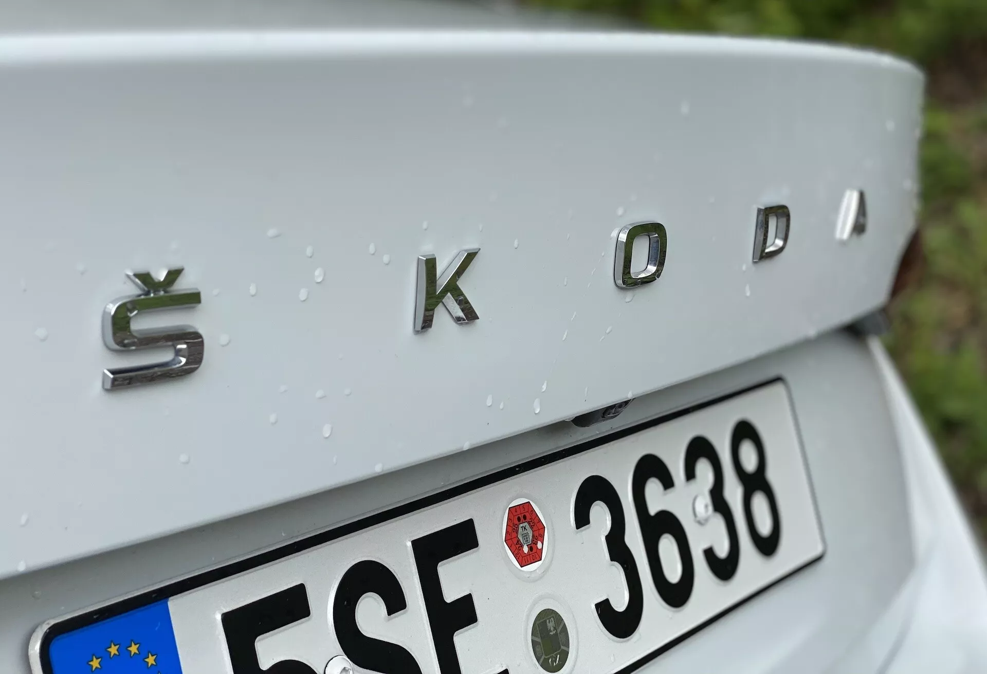 Közelkép egy 2020-as Škoda Octavia (octavia-limousine) hátsó részéről, a háromdimenziós fémes kivitelű 'ŠKODA' krómozott logóval a középpontban. A felvétel kissé felülről lefelé mutatja a finoman lejtő csomagtérfedelét, részben látható európai stílusú rendszámtáblával alatta, és esőcseppekkel díszített ezüst autófelülettel, jelezve a nemrég lezúdult esőt.