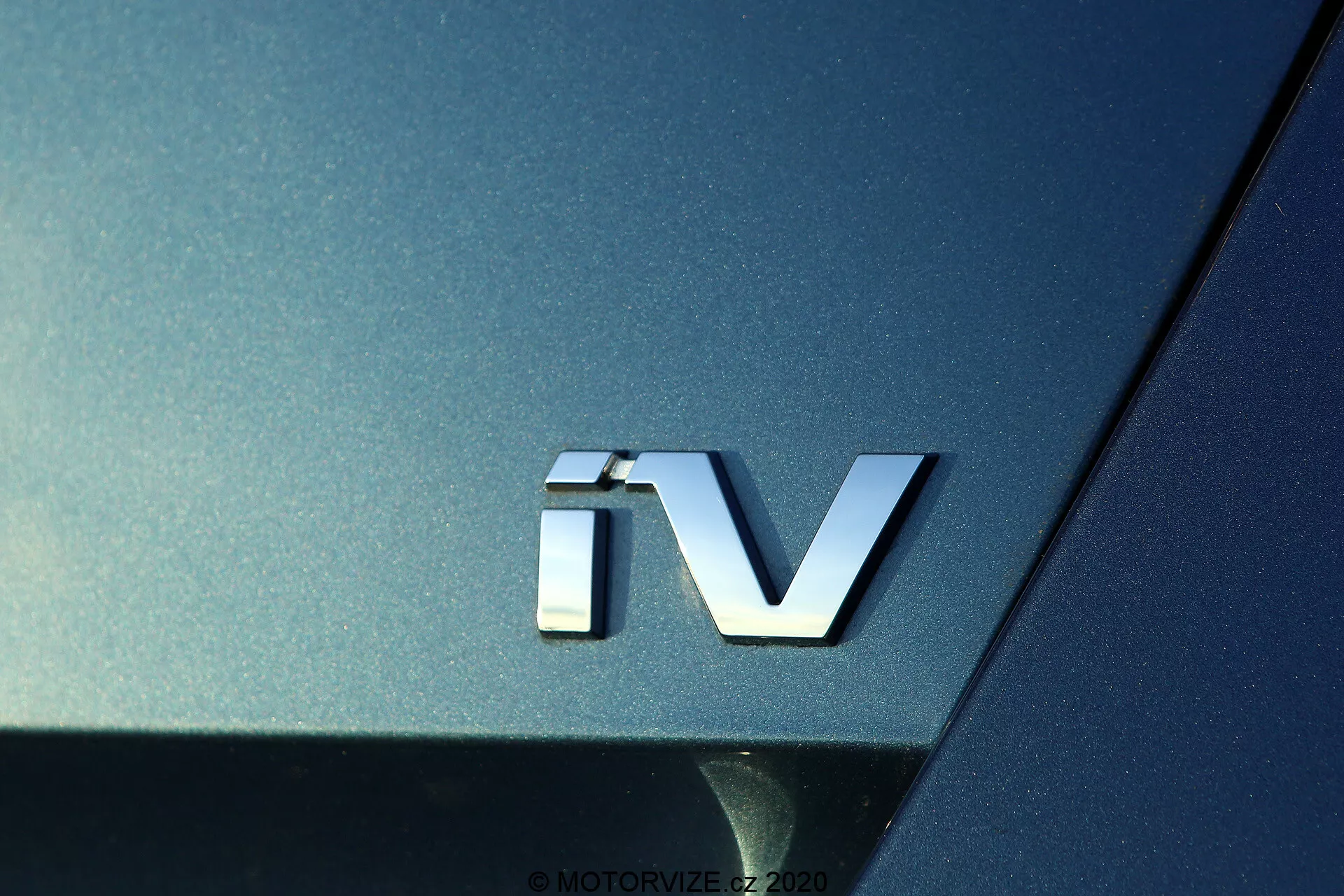 2020-as Skoda Octavia 'iV' jelzésének közeli képe, amely elektromos hajtásláncot jelöl, ezüst 3D emblémával a sötét fémes karosszérián, kiemelve az autó hátsó szakaszának ívelt felületét és visszaverő tulajdonságait.