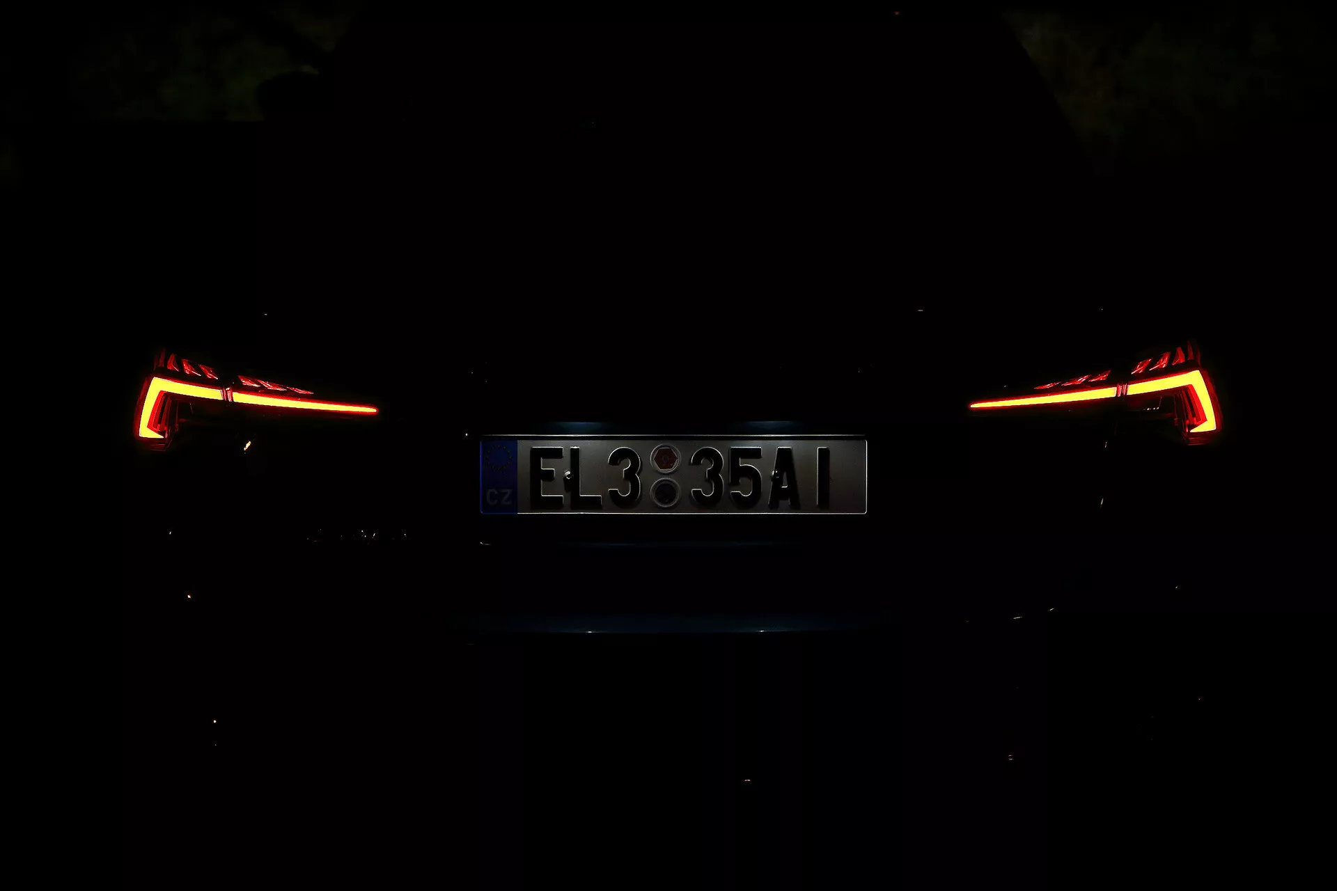 Skoda Octavia 2020 Limousine hátsó nézete, éjszaka vagy gyengén megvilágított környezetben fényképezve. A kép a megvilágított hátsó lámpákra és rendszámtáblára összpontosít, modern és szögletes kialakítást mutatva. Az autó részletei gyenge megvilágítás miatt homályosak, kiemelve az elegáns és korszerű hátsó részt.